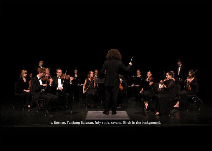 A video still depicts musicians in black dress clothes playing instruments in an orchestra, a conductor stands in the centre facing away from the camera. White subtitles line the bottom of the frame.