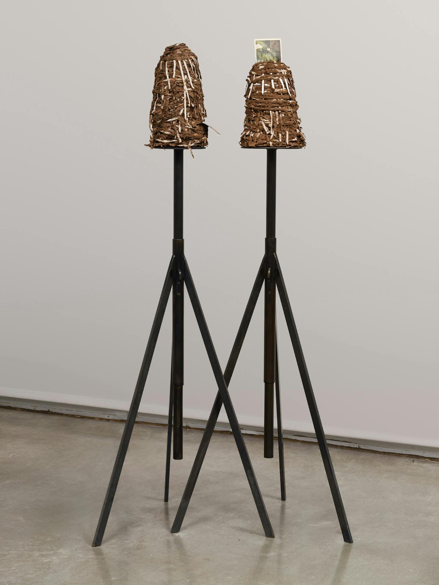 A sculpture in a gallery space. The sculpture consists of two moud-like forms made of brown adobe mud and strips of tape, each sitting atop a metal, tripod-like form.