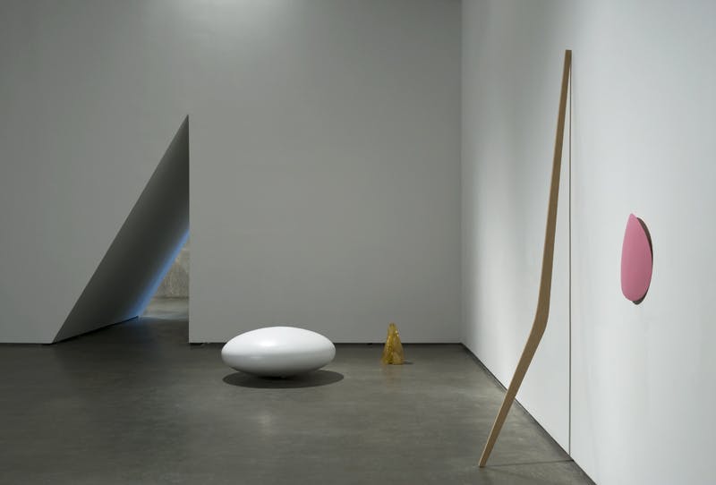 A white, oval stone-like sculpture is next to the entrance of a gallery. A wooden, bow-like sculpture leans against the white wall next to a pink oval sculpture, slightly bent and mounted on the wall.