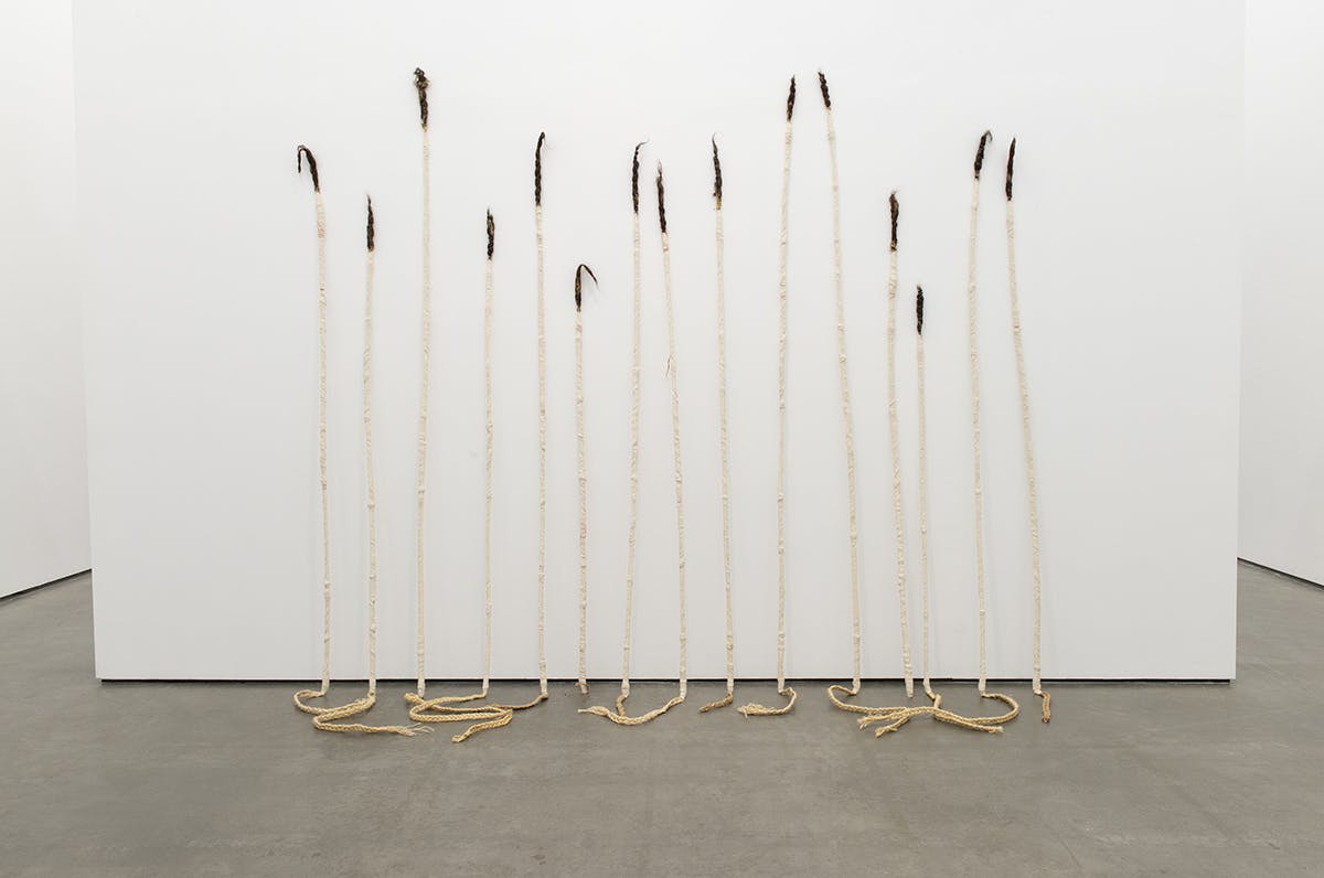 A set of fifteen tall, thin sculptures are leaning against a gallery wall. Their bodies are made of braided grass wrapped in white cotton. Human hair emerges from the top of each sculpture.