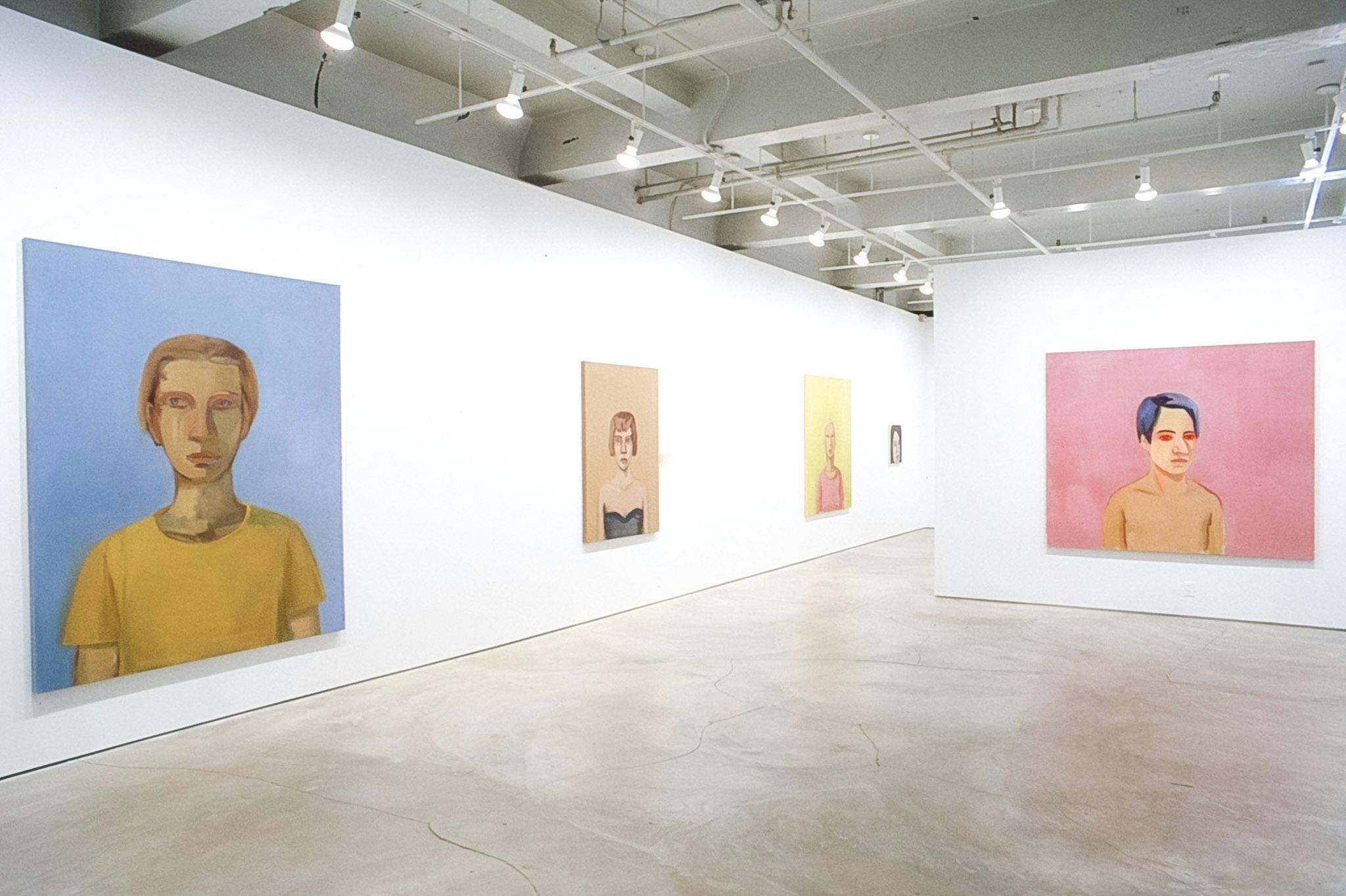 Five portrait paintings are visible in this photograph. The large-sized painting on the left depicts a man in a yellow T-shirt standing in a blue background. His eyes are looking away. 