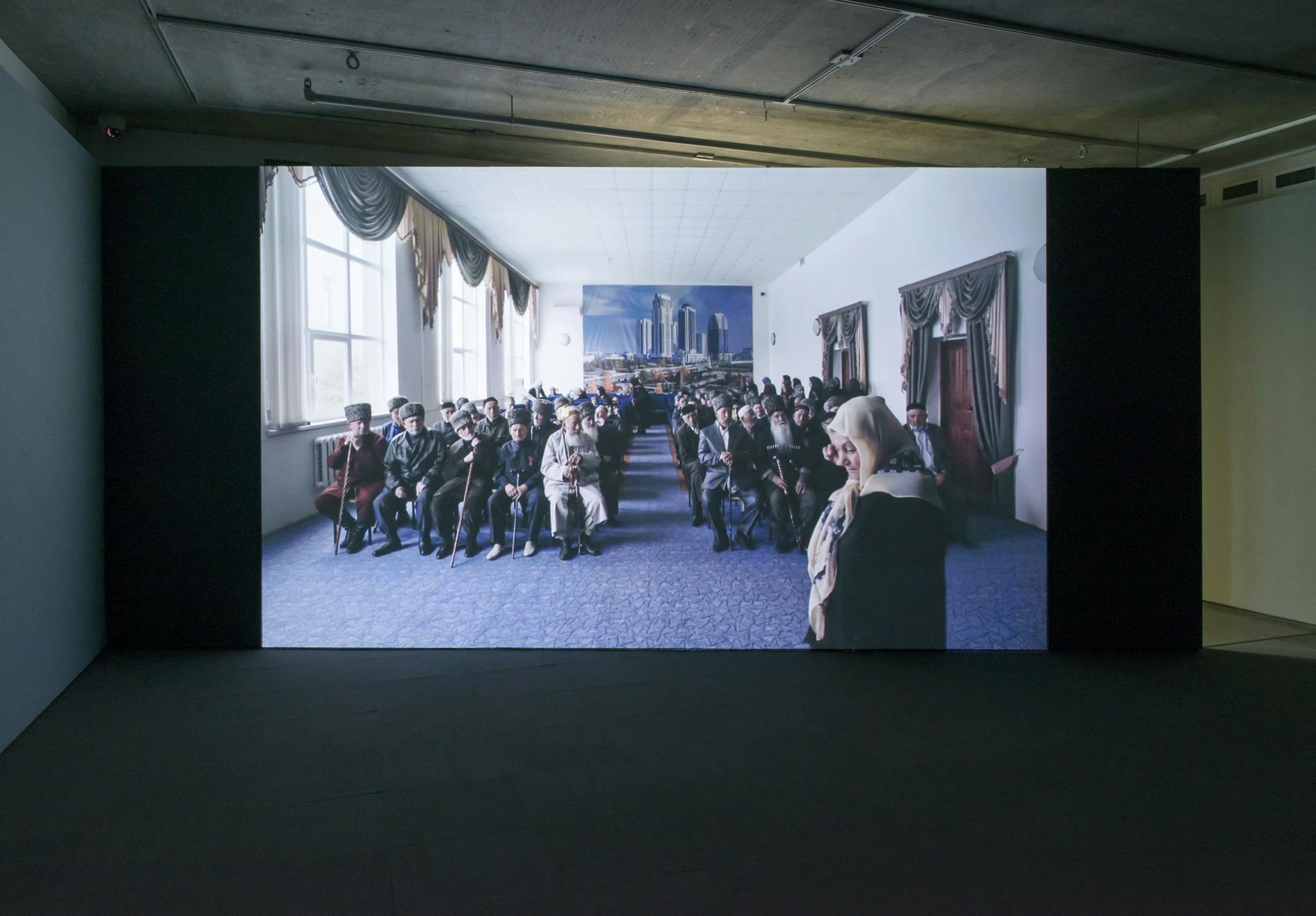 A large screen is displayed in a dark gallery. On the screen, a group of people sit facing the front of a large room. A person stands at the front of the room with their face turned away.