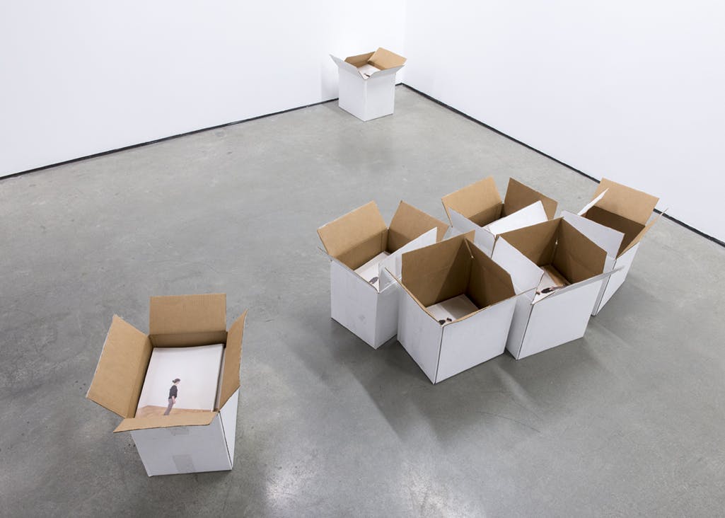 Seven white cardboard boxes are grouped on a gallery floor. The boxes are open and filled with the publications for the exhibition. The cover of the books are visible.
