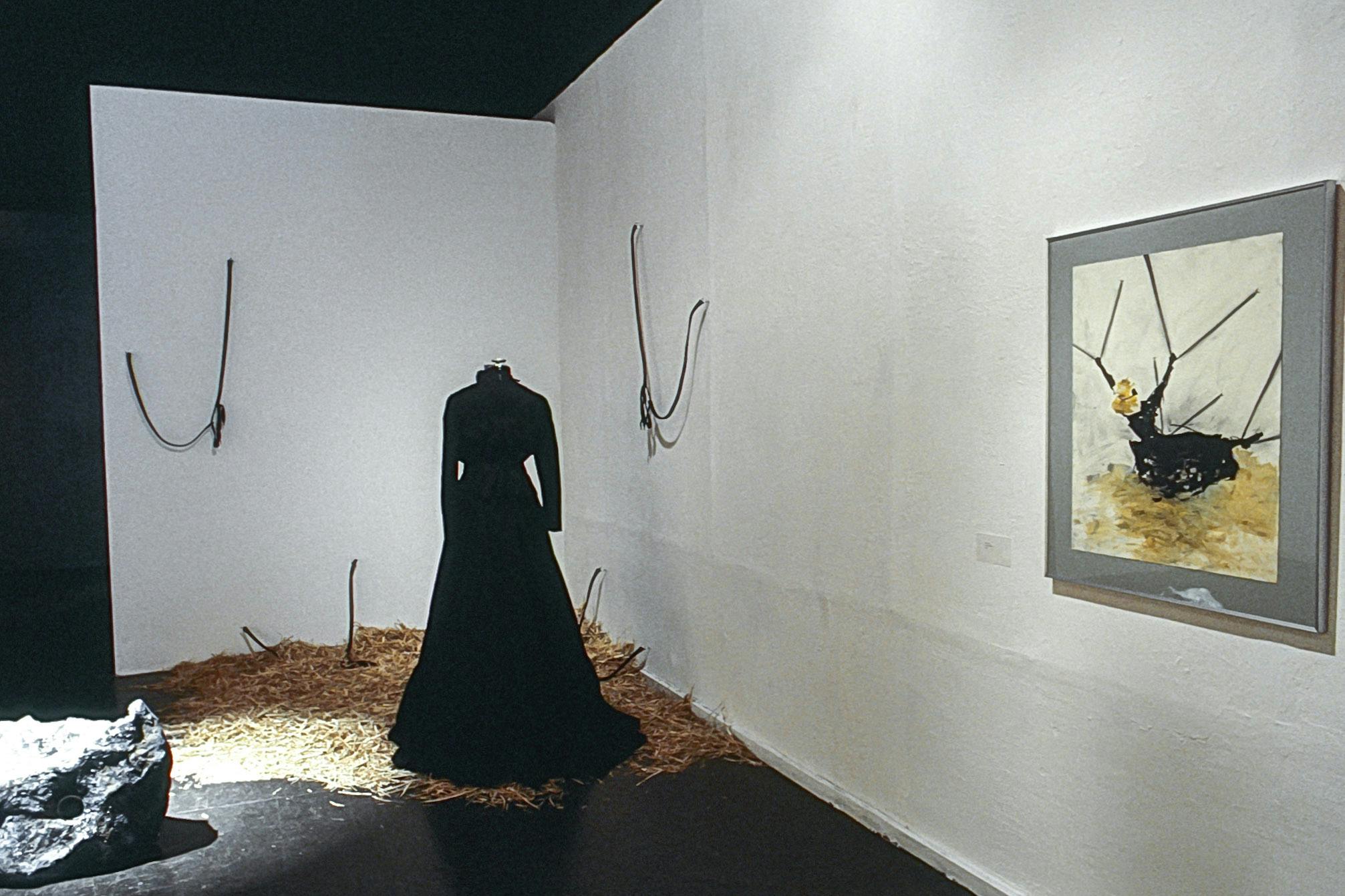 In the corner of a gallery, there is a long black dress on a mannequin, resting on pile of hay on the floor. Several cables are in the hay and on the wall, as well as a rock sculpture and a painting.