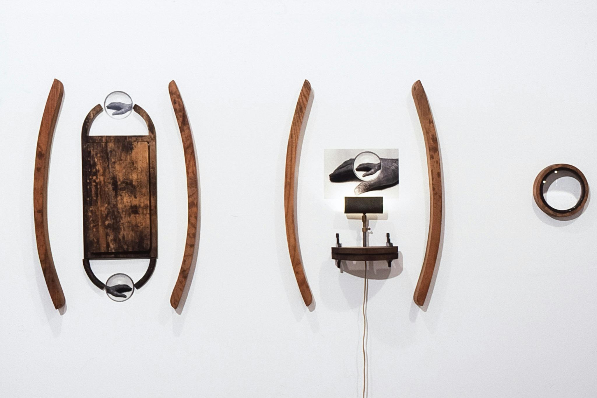 Three pieces of sculptures are mounted on the wall. Wooden pieces including a cutting board, monocular lenses, and the identical image of a hand are repeated throughout the composition.