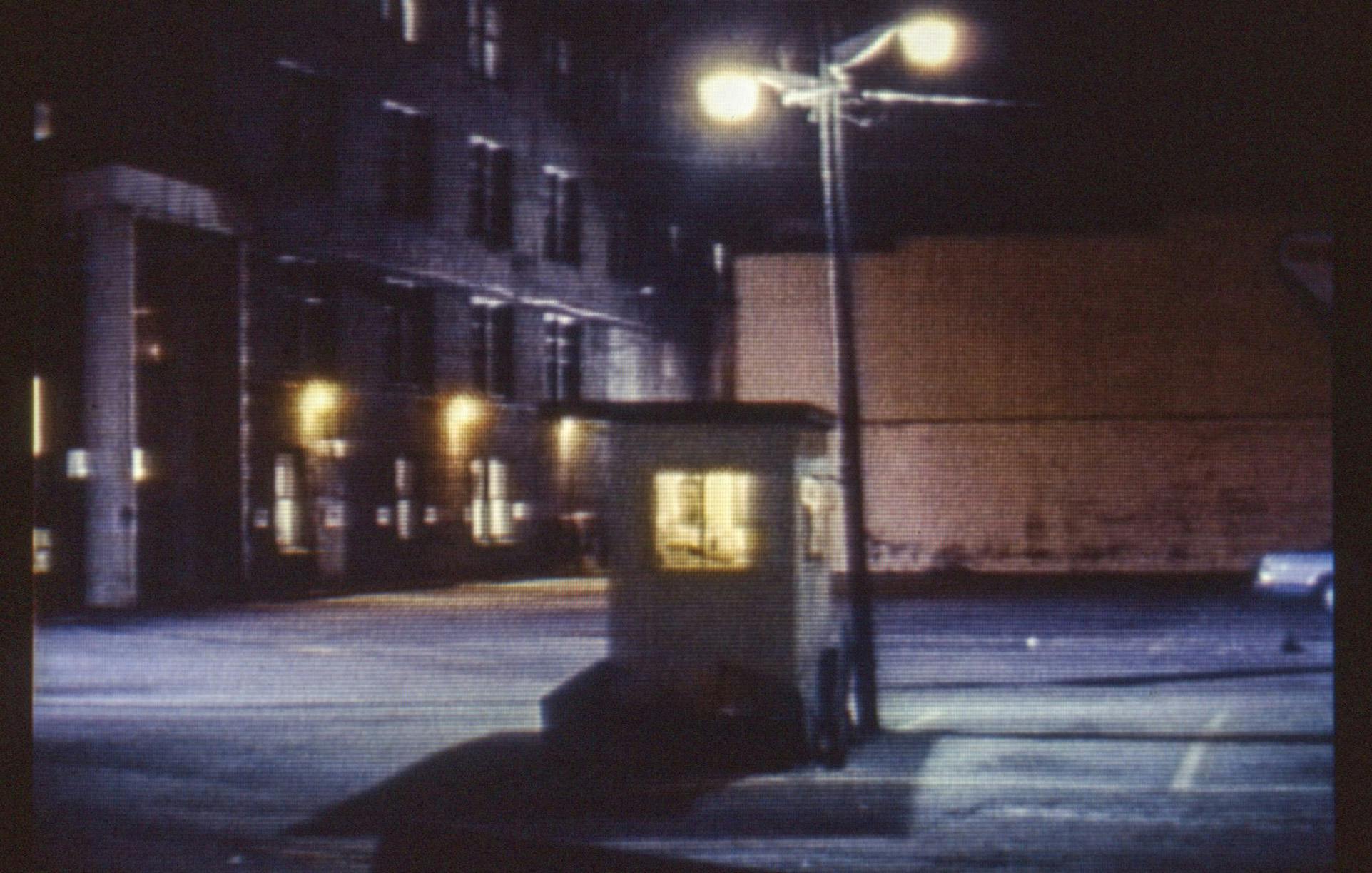 A still image from a film on a small screen. The photo is a closeup, the grainy texture of the film is visible. The scene shown is an empty outdoor parking lot at night, with a person at the pay booth.