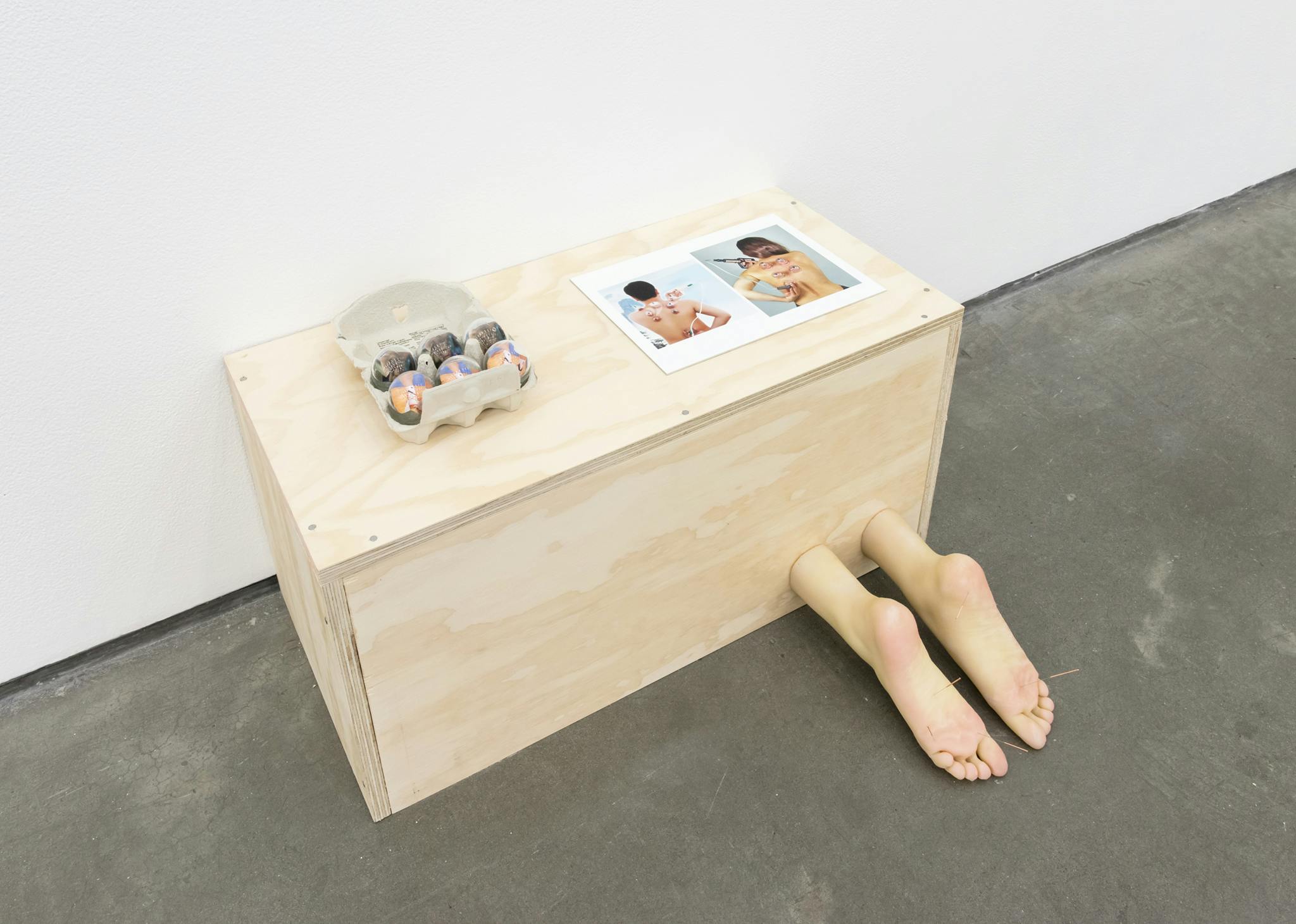 A sculpture sits on the floor against a wall of a gallery. The sculpture is a wooden box with prosthetic feet protruding out in front. Two photographs of people’s backs and an egg carton sit on top. 
