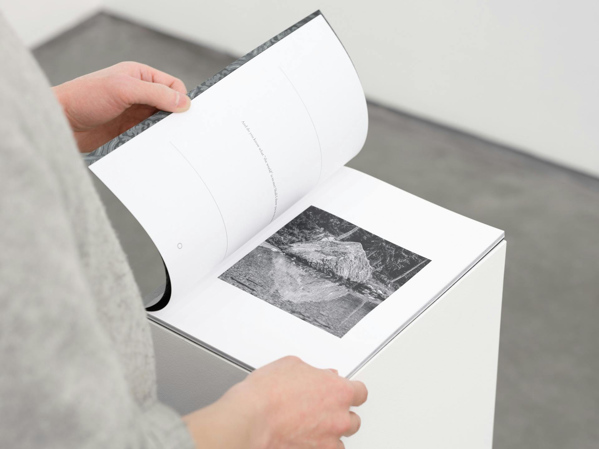 A thin artist book on a white plinth. A pair of hands hold a page open to show a black and white photograph of a large rock and its reflection in a lake.