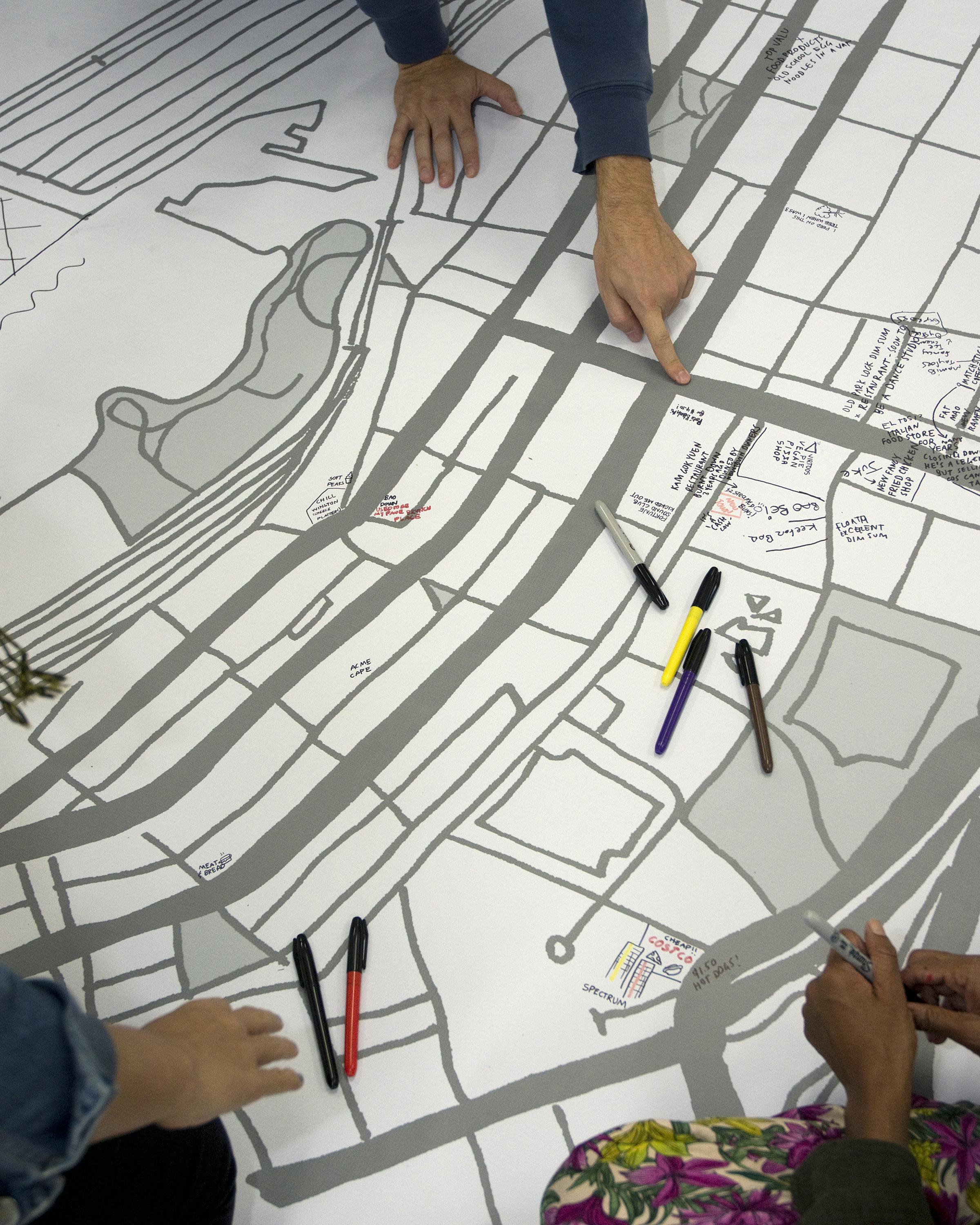 A large black and white map covers the floor. Various coloured markers are scattered across the surface and a person is pointing to an intersection on the map. 