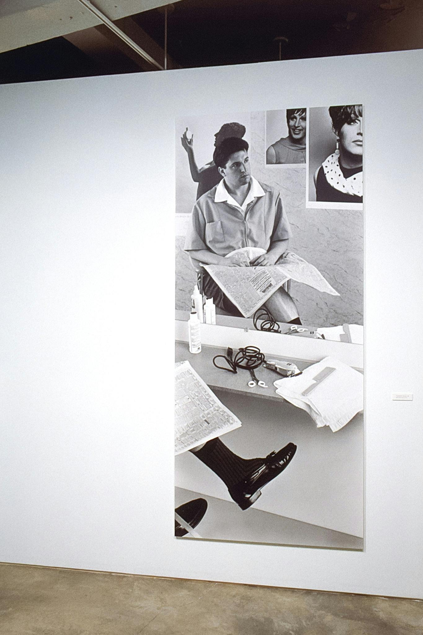 A black and white photograph is installed on the gallery wall. In this tall rectangular-shaped photo, a man sits on a chair in front of a large mirror. He looks away from the newspaper on his lap.  