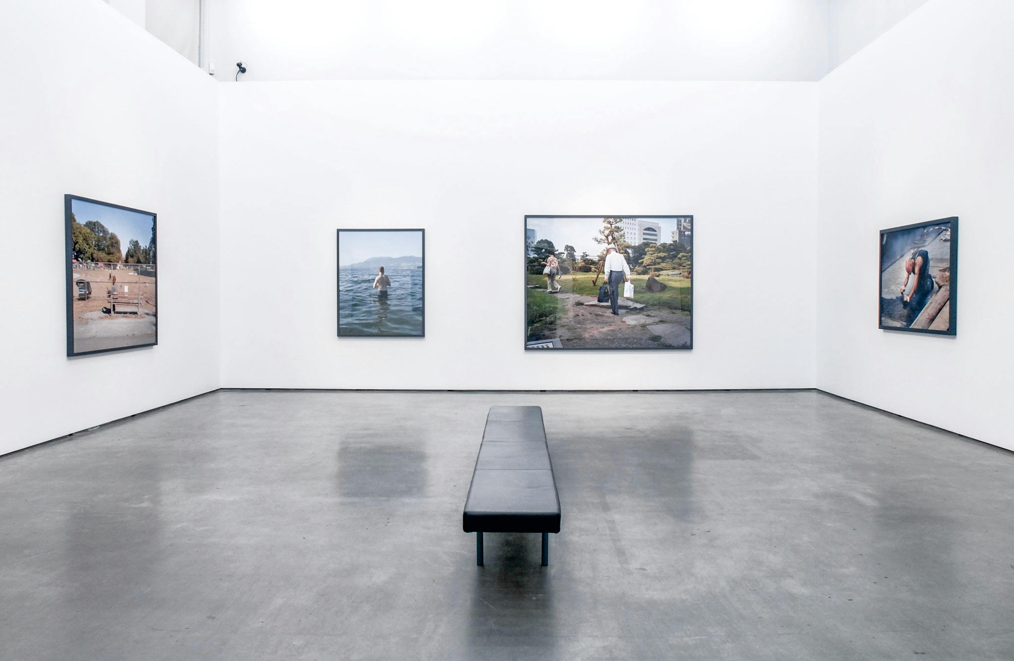 Four large-sized photographs are installed on the gallery walls. One photograph depicts a person standing in a body of water. The other photograph captures two people walking in a park. 