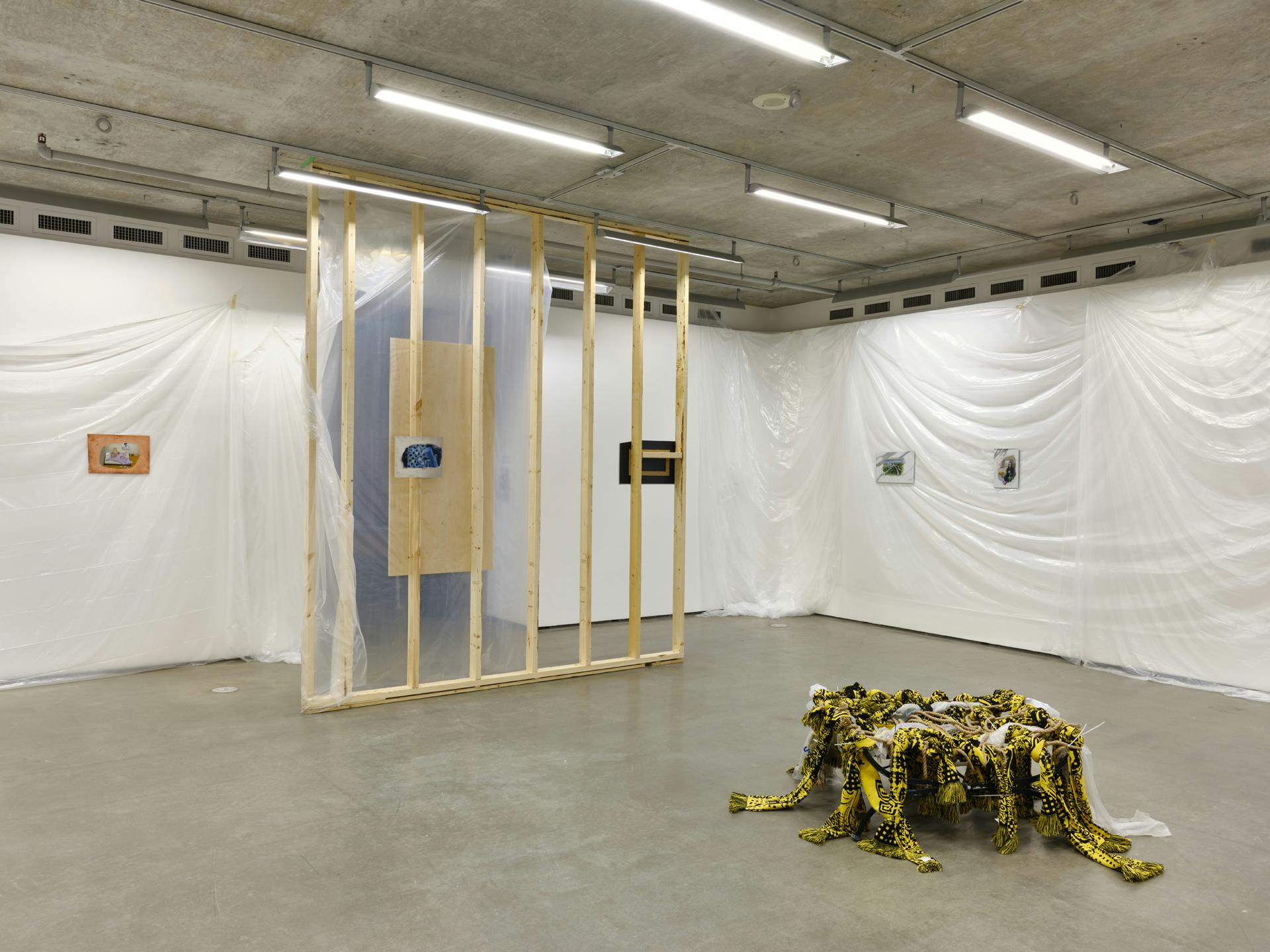 Small paintings hang on gallery walls draped with plastic sheets. At the entrance is an unfinished wall structure, paintings hang on either side of it. A sculpture made of textiles, plastic and rope sits on the floor. 