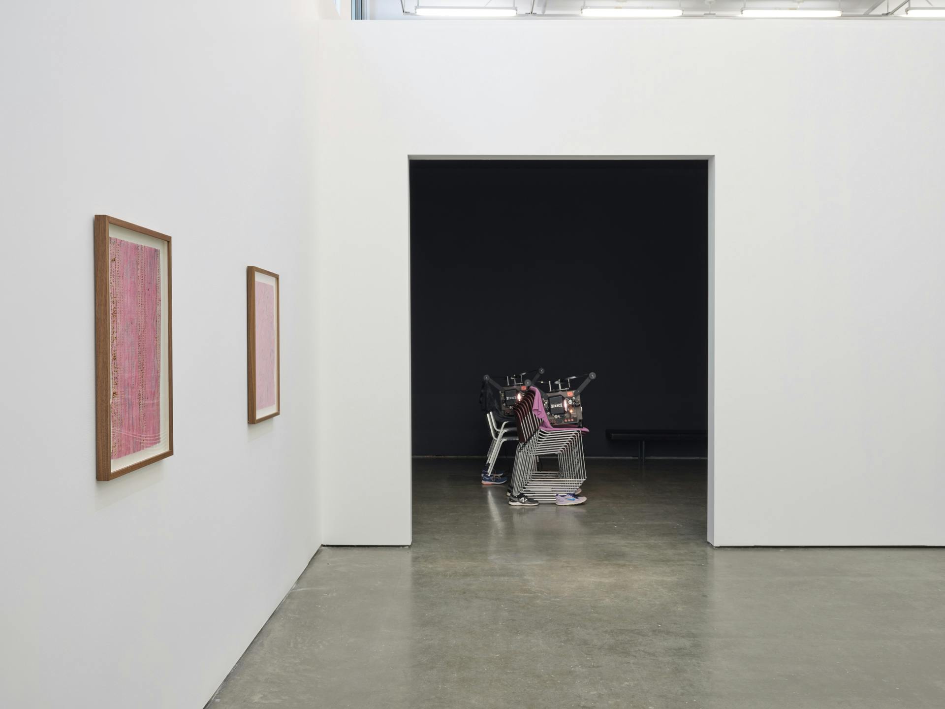 Two framed pink collages on a white wall perpendicular to a doorway leading into a dark room with a sculpture made of stacked chairs supporting a film projector visible.