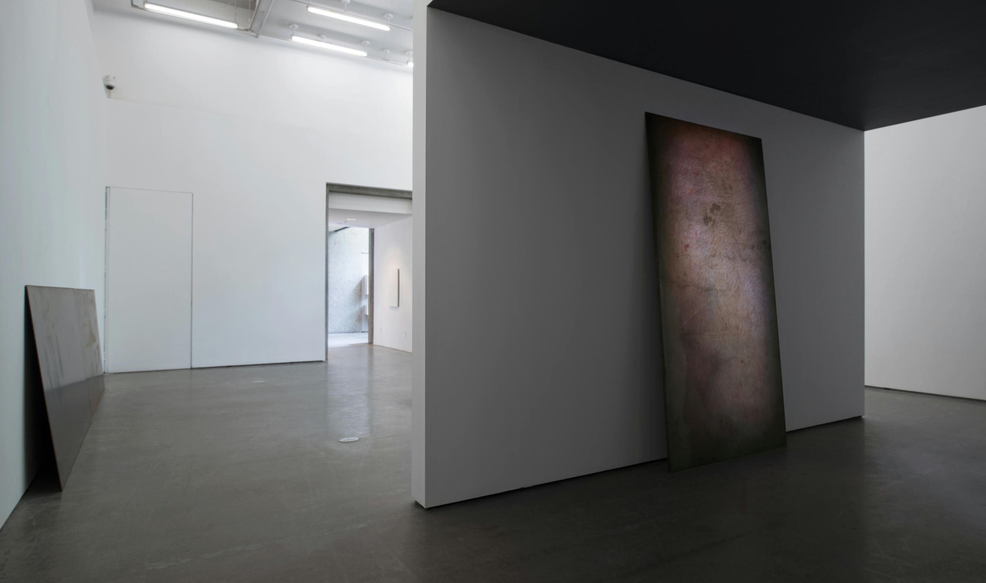At the center of a gallery space, one large steel sheet leans against the back of a freestanding wall. A low ceiling darkens the space around the sheet and wall. 