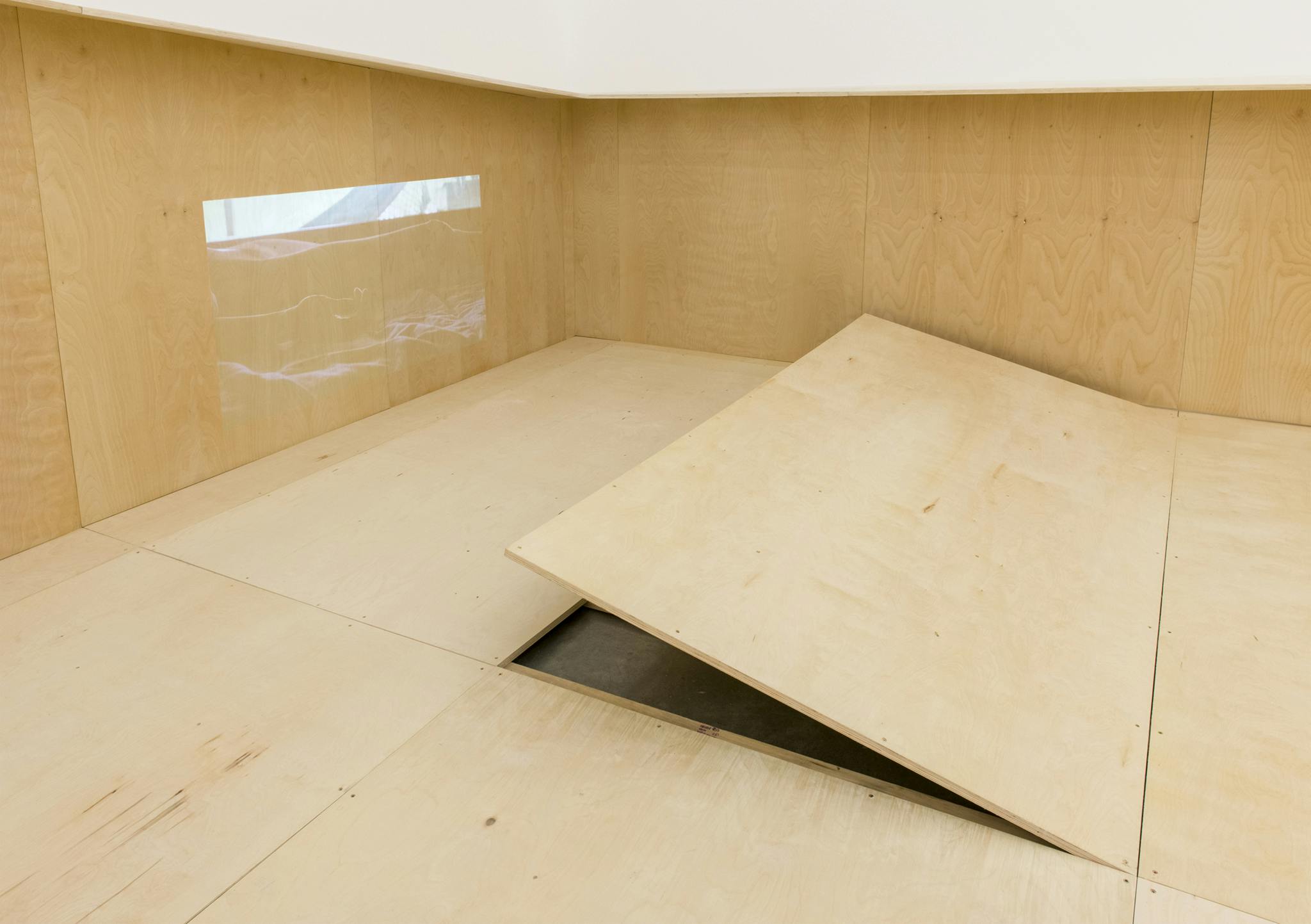 In the corner of a gallery, one of the large rectangular plywood floor panels is partially lifted up, revealing a bit of the concrete floor below. A projected image is cast on the wall opposite. 
