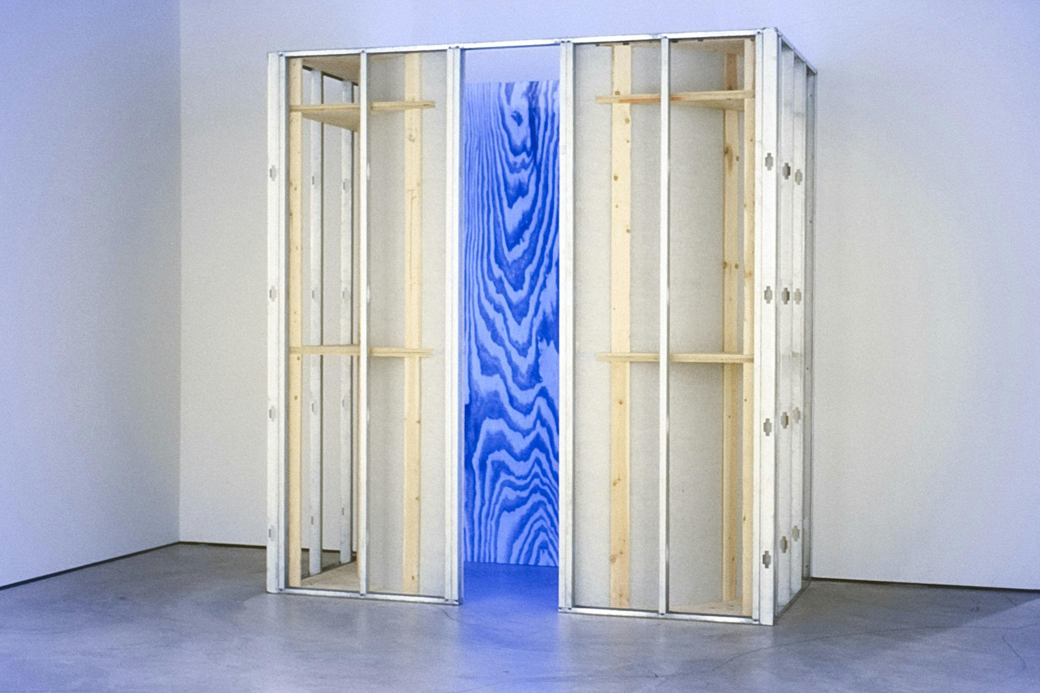 A large sculpture in a gallery space. It is made of a wood and metal structure and shows an enlarged wood pattern in the interior. This inside part is lit in bright-blue.