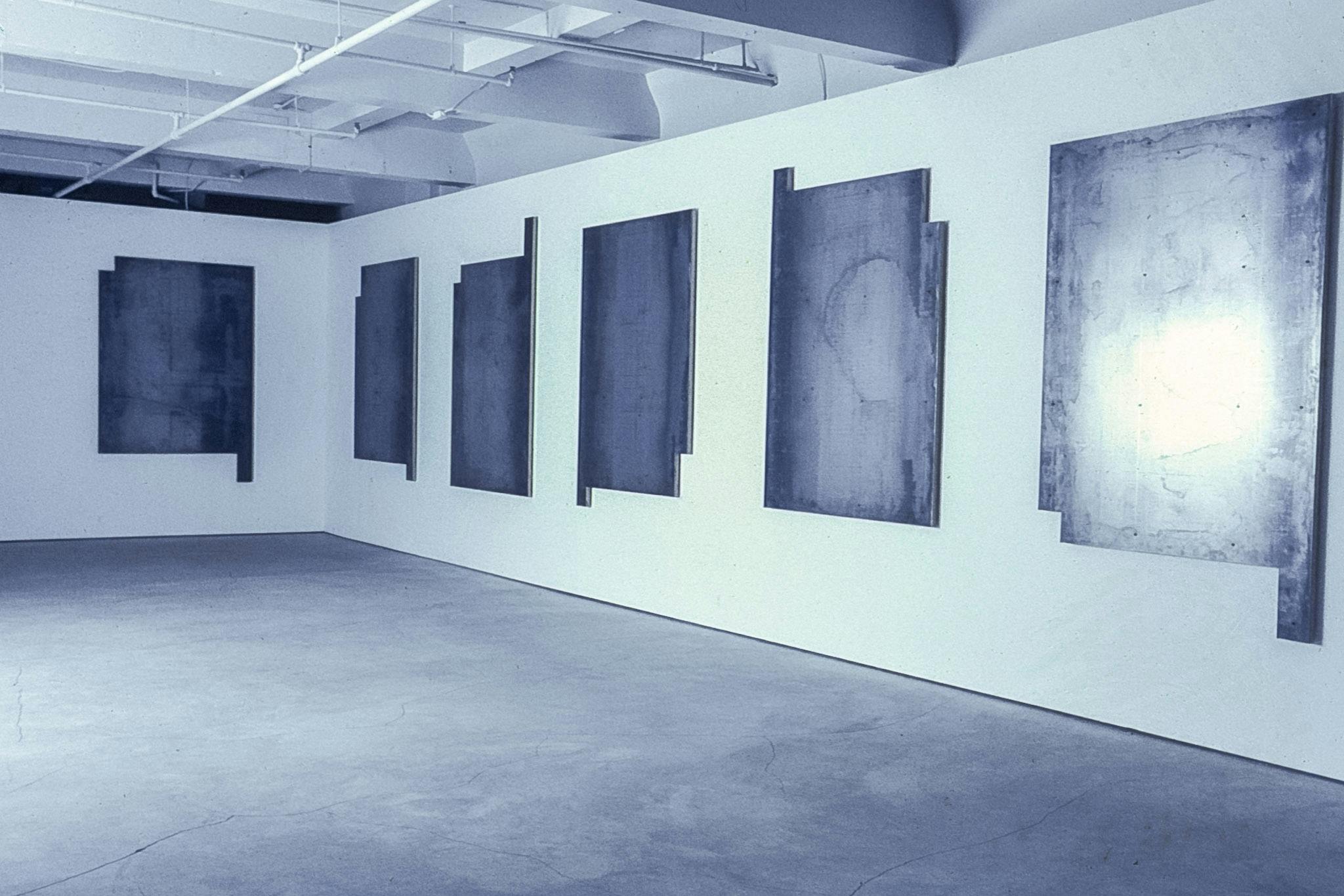 In the dark corner of a gallery, several rectangular metal panels hang on the wall. Their surfaces are scuffed and marked, and the single light source in the room reflects brightly from them.