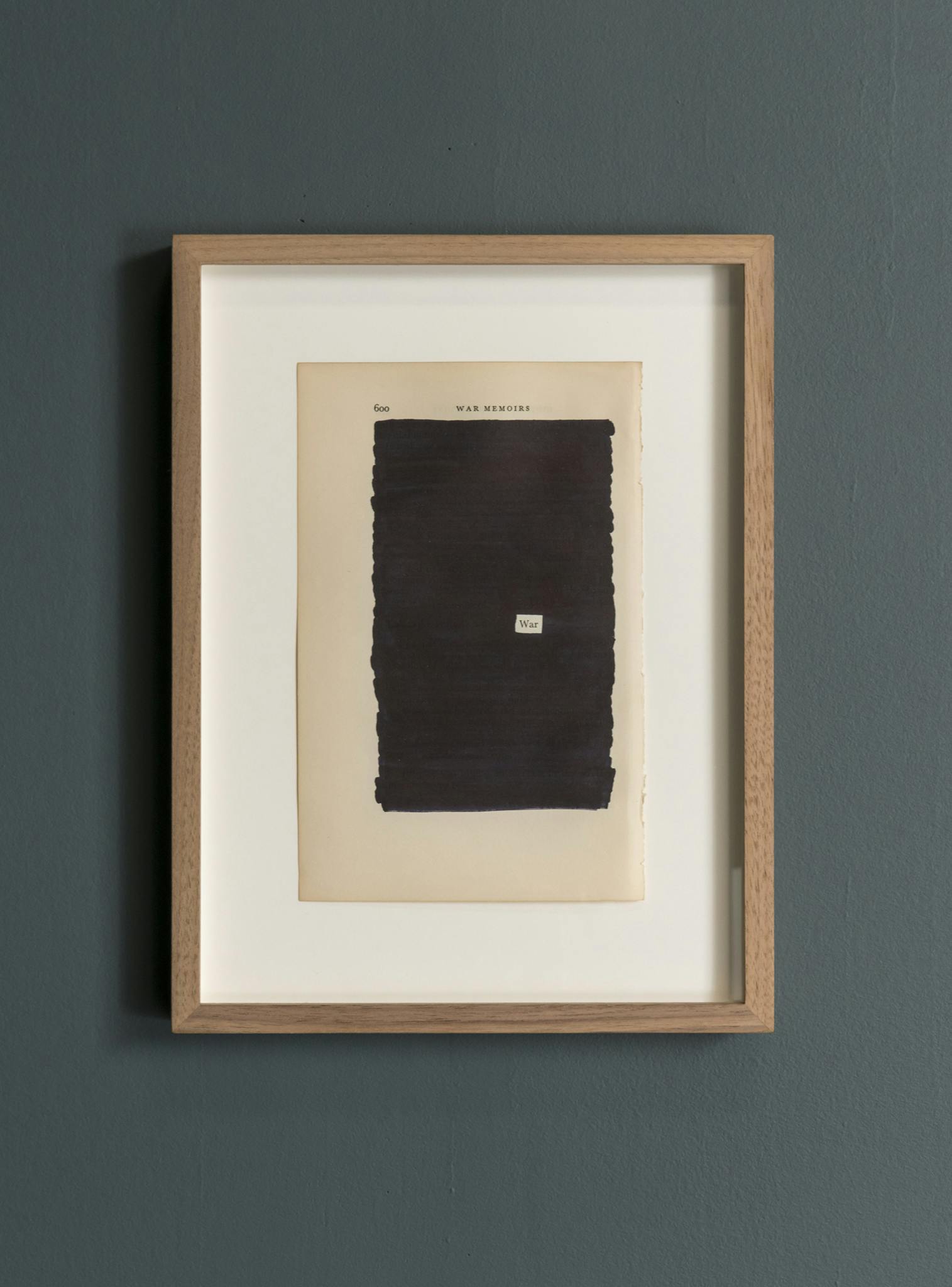 A wooden frame hangs on a blue-grey wall. Mounted in the frame is a page from a book. The title of the page reads “WAR MEMORIES.” The rest of the page is redacted, excluding one word, “war.” 
