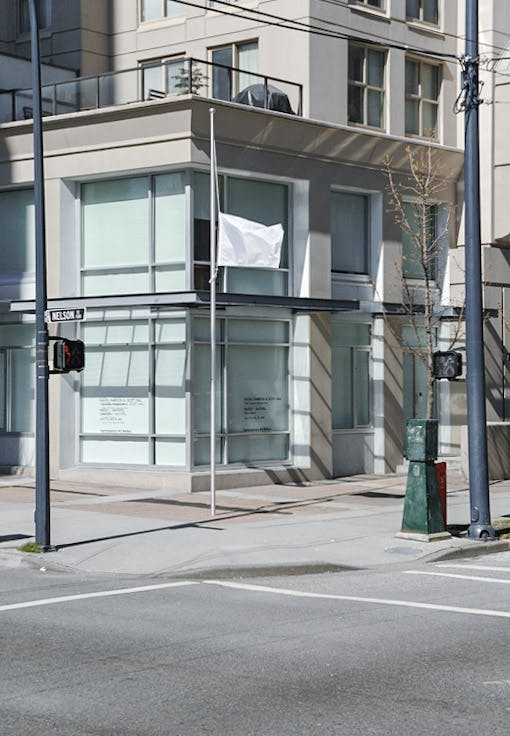 A flagpole is erected on a sidewalk in front of CAG. A white flag is half-masted on that pole. The pole is about the same height as the two-story building. 
