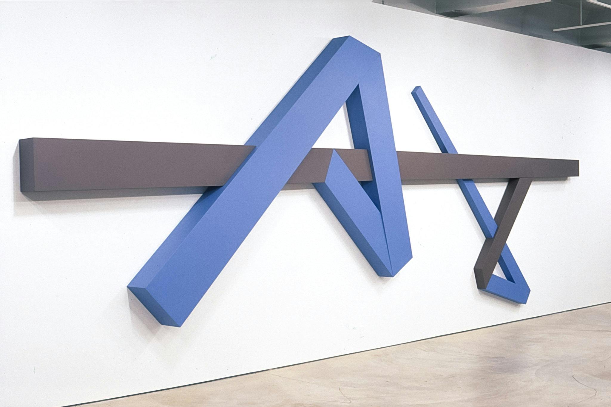 A large-sized geometric sculpture is installed on the gallery wall. Two blue incomplete arrow shapes are connected by the brown straight line that runs from the left to the right of the wall. 