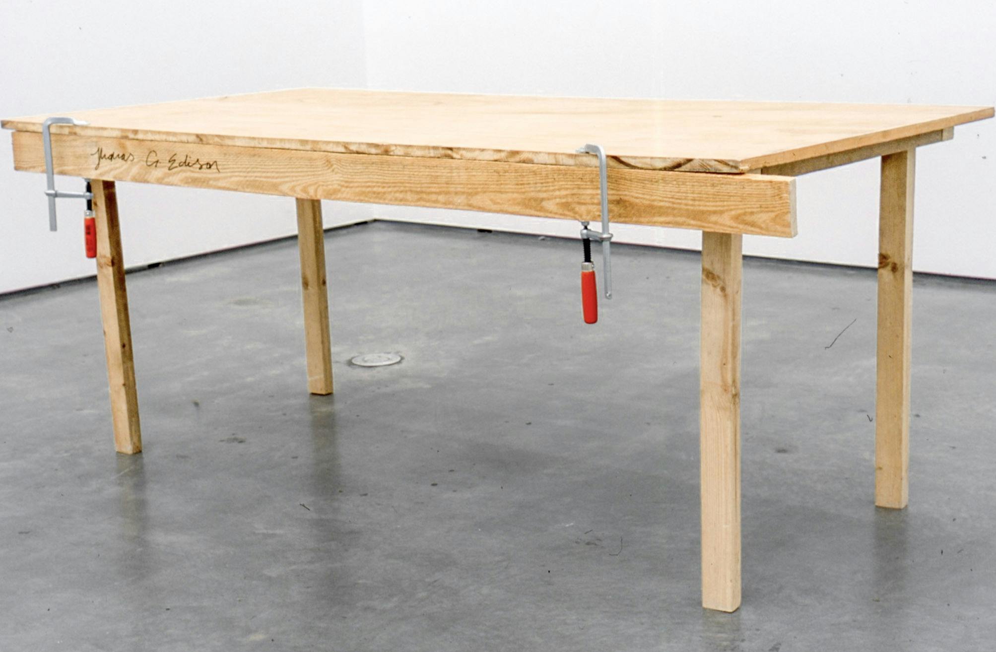 Detail of an installation work by KirstenPieroth. A wood table placed on a gallery floor has a signature that is read “Thomas G. Edison.” Two bar clamps are attached to the long side of the table.