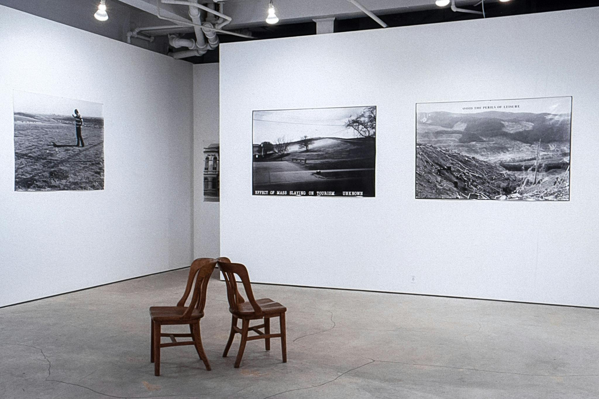 A gallery with large photos on a wall. One shows an empty park, with text that reads "Effect of Mass Slaying on Tourism, Unknown." In the center of the room, two wood chairs sit back to back. 