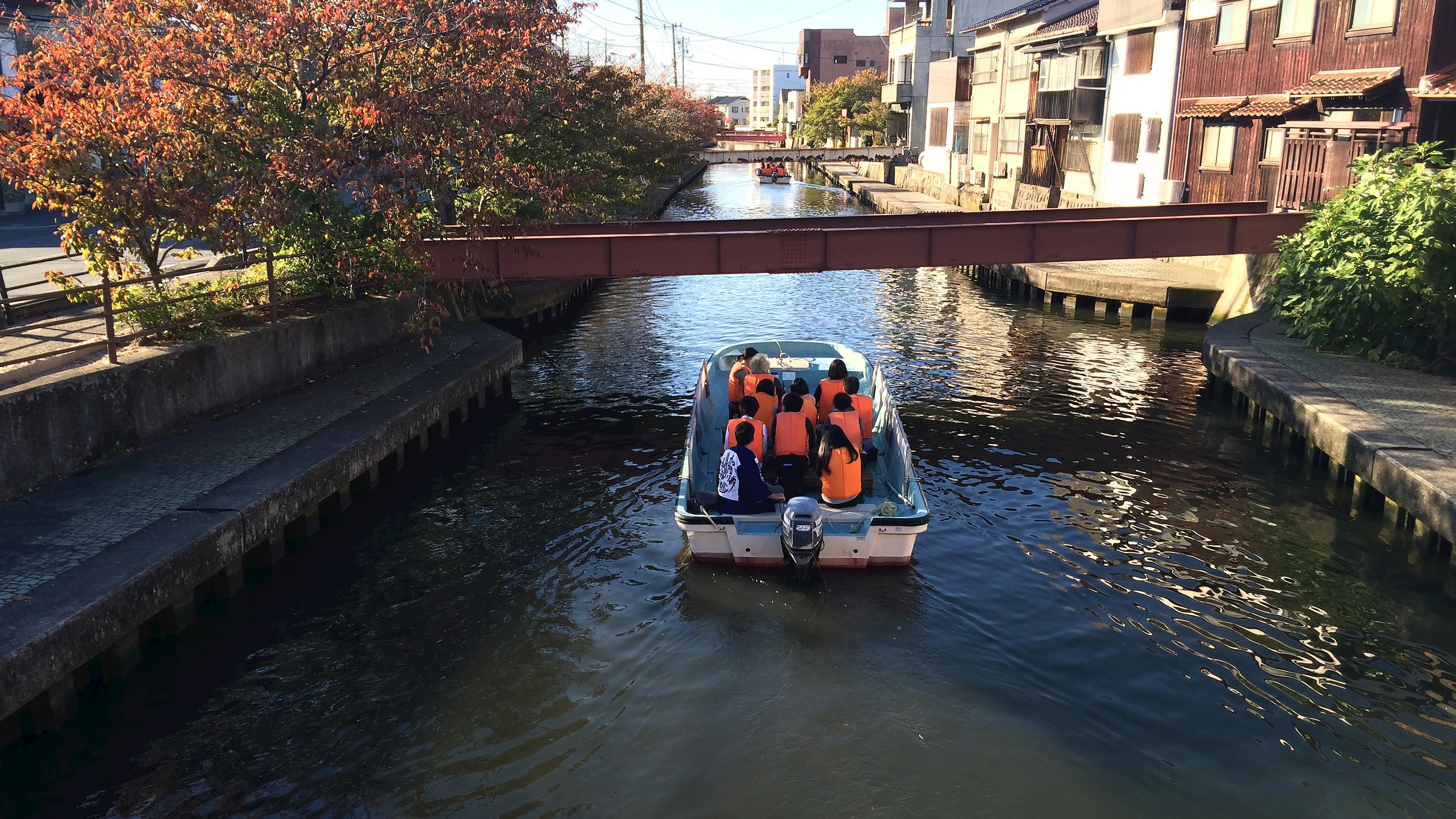 An erial view of a small white boat on a river that flows through the village. Ten people wearing orange life jackets and a person in a blue happi coat sit on the boat facing the front.