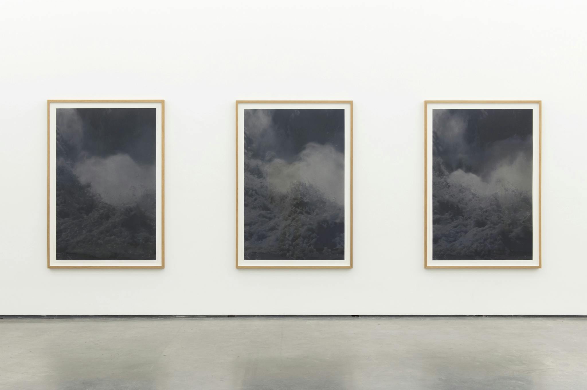 Three large-scale framed photographs hang on the wall of a gallery space. Frothing water is visible in the low-contrast, blue-grey toned images. 