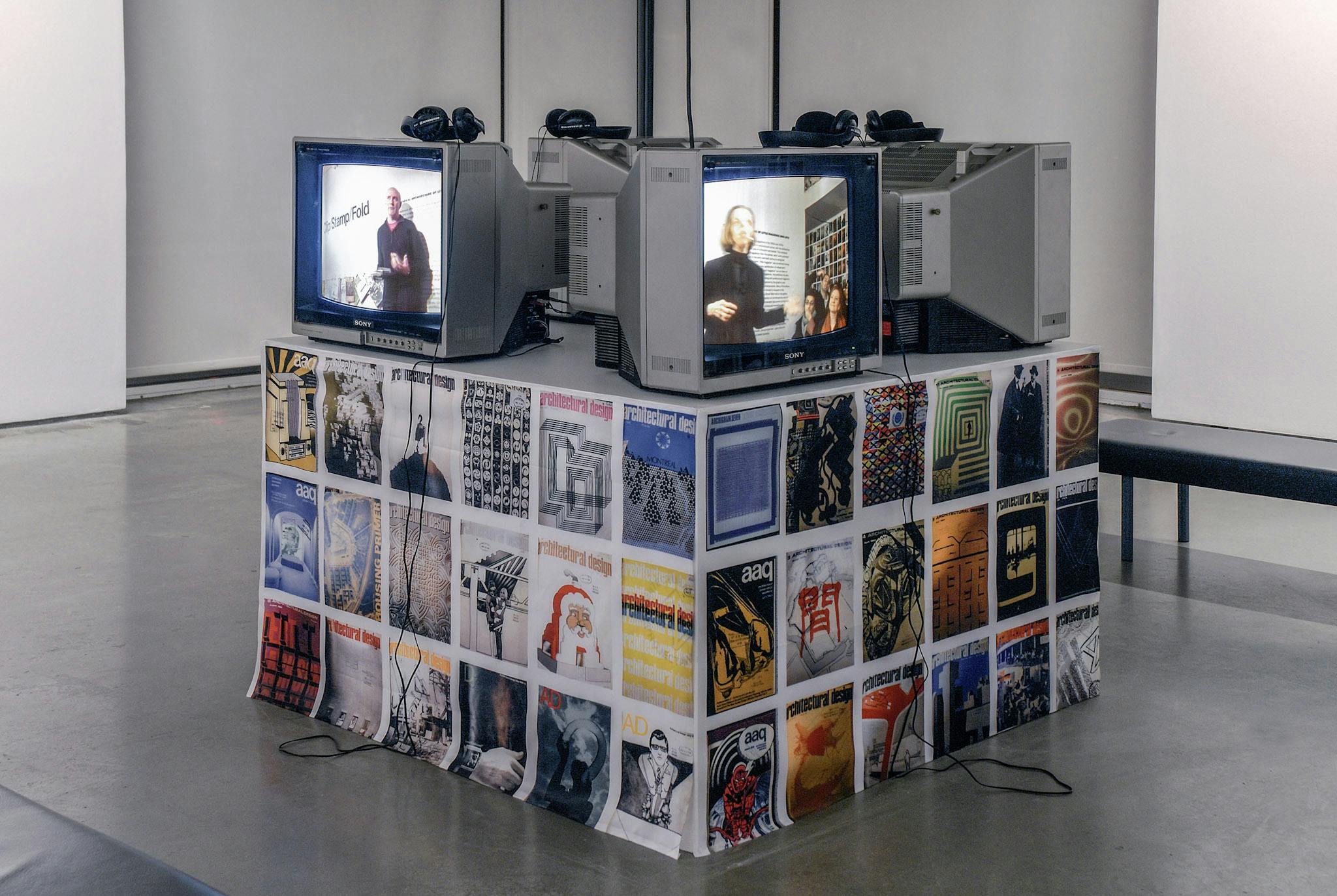 Four CRT TVs are placed on a large pedestal. They play different videos of a person giving a speech in public. The pedestal is covered by the printed images of various magazine covers.