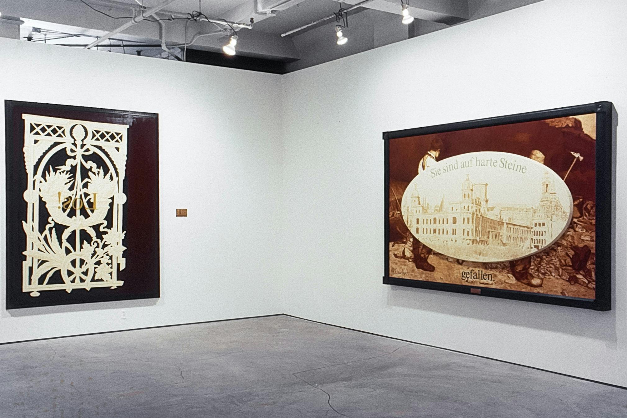 2 large paintings in the corner of a gallery. The one on the left shows an ornate gate pattern and backwards yellow text reading "Los!" The one on the right shows a building, workers, and German text.