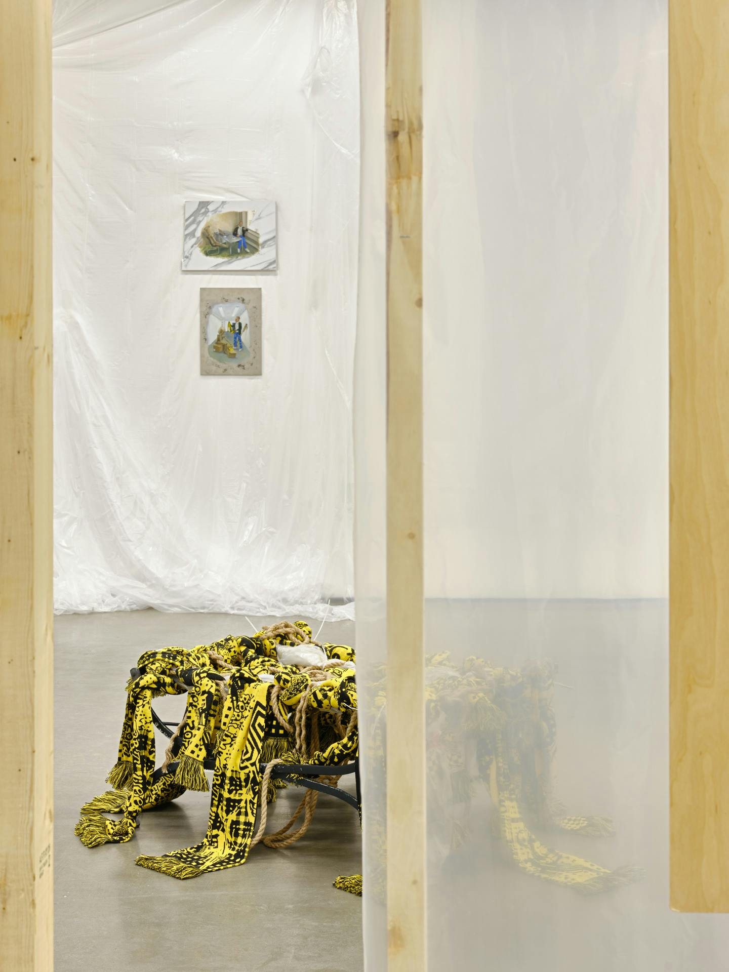 A view through an unfinished wooden wall structure of two small paintings hanging on a gallery wall draped with translucent plastic sheets and a sculpture sitting on the floor made with yellow textiles. 
