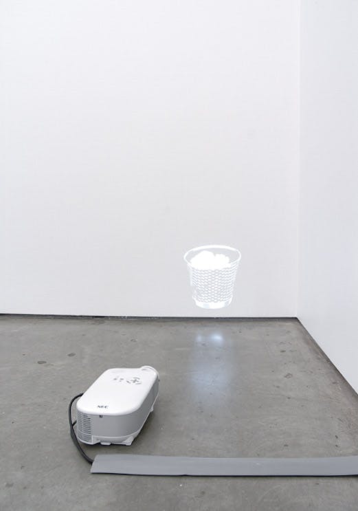 An installation view of a work titled Trash by Ceal Floyer. A small projector is placed directly on the floor. It projects an image of a trash can on the wall close to the corner of a gallery.