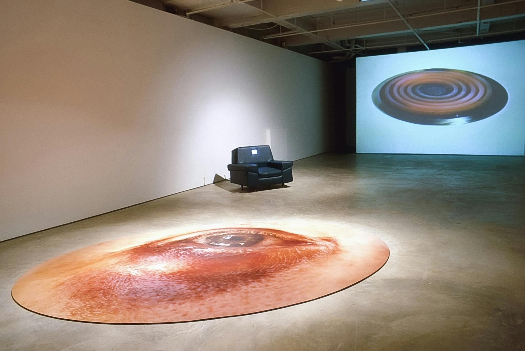 Three artworks are installed in the gallery. A projection of a coil stove is on the wall, foregrounded by a black rug chair on the floor. A circular-shaped photograph of an eye sits beside the rug. 