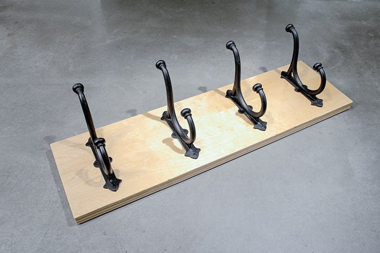 A coat rack is placed flat on the floor. It is a set of four black coat hooks mounted on a rectangular piece of wood.