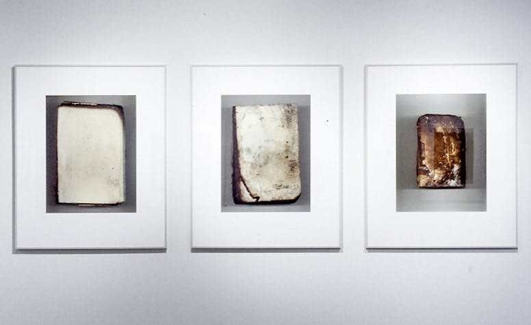 A closeup of 3 vertical photos in silver frames, mounted on a white wall. The images are of covers of books, indecipherable due to fire damage with dark and burnt edges, crinkled and textured.
