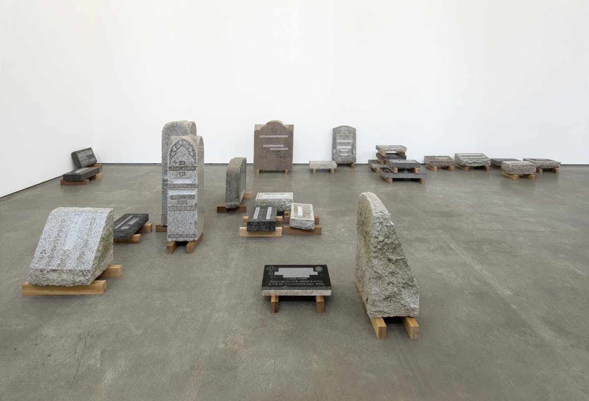 Variously sized tombstones installed in a gallery. Some of them are a pink stone, while the rest are black, grey, or white. Each stone sits on a pair of wooden slats.