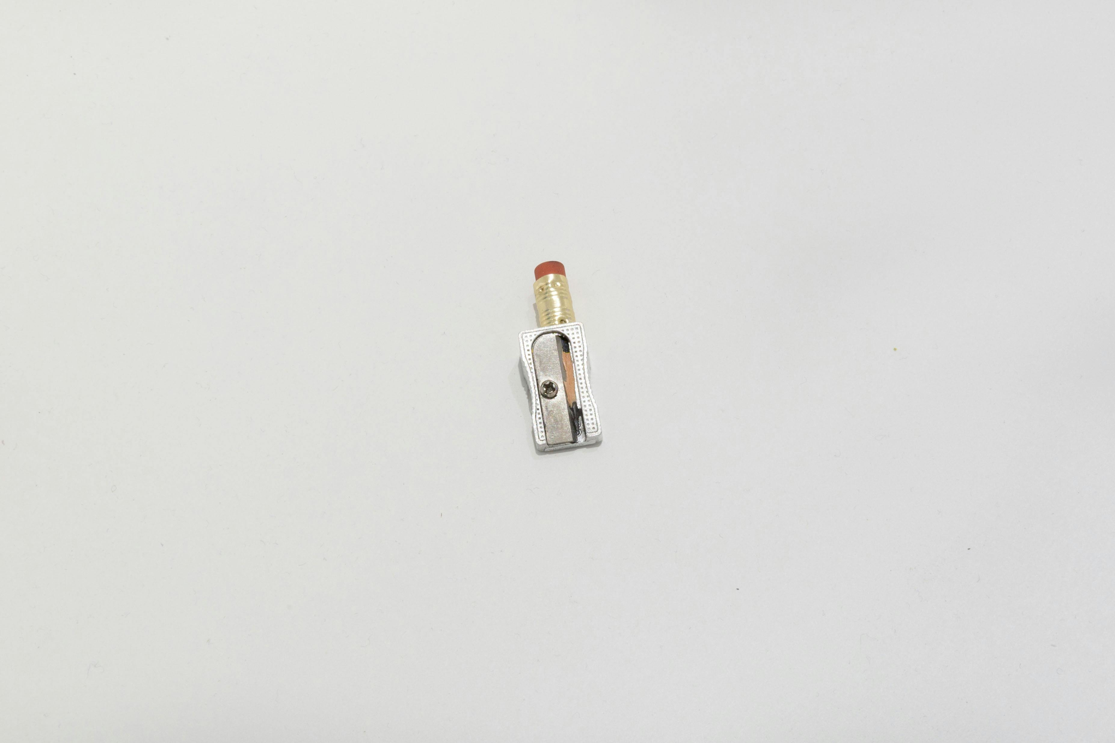 A silver pencil sharpener is placed on a white flat surface. The tip of a black pencil, sharpened down to the pink eraser end, is inserted into the sharpener. 