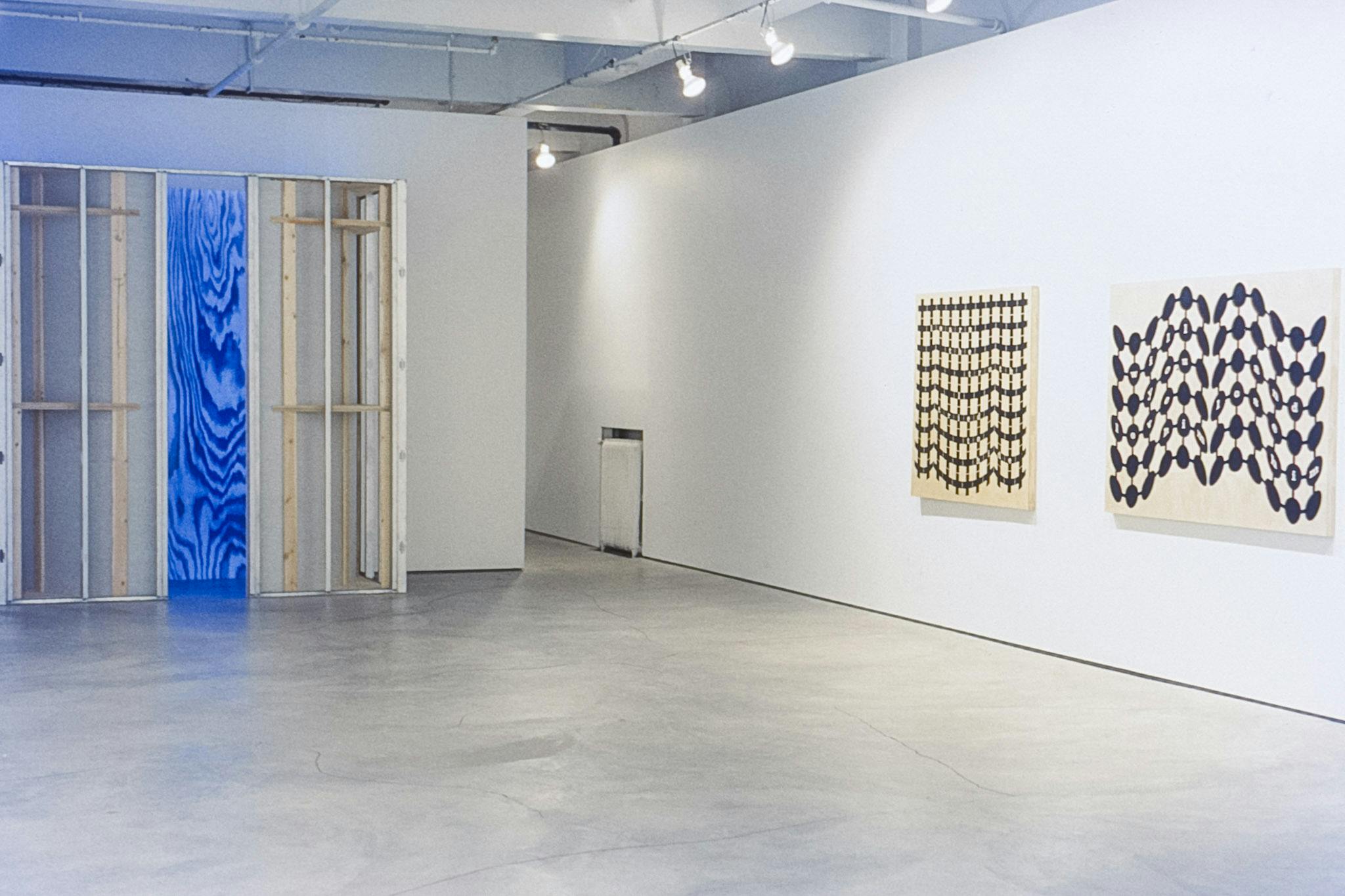 The corner of a gallery space with 3 artworks visible. One is a wooden structure that shows a large wood pattern inside, in blue. The other 2 works are wood panels with black geometric patterns. 