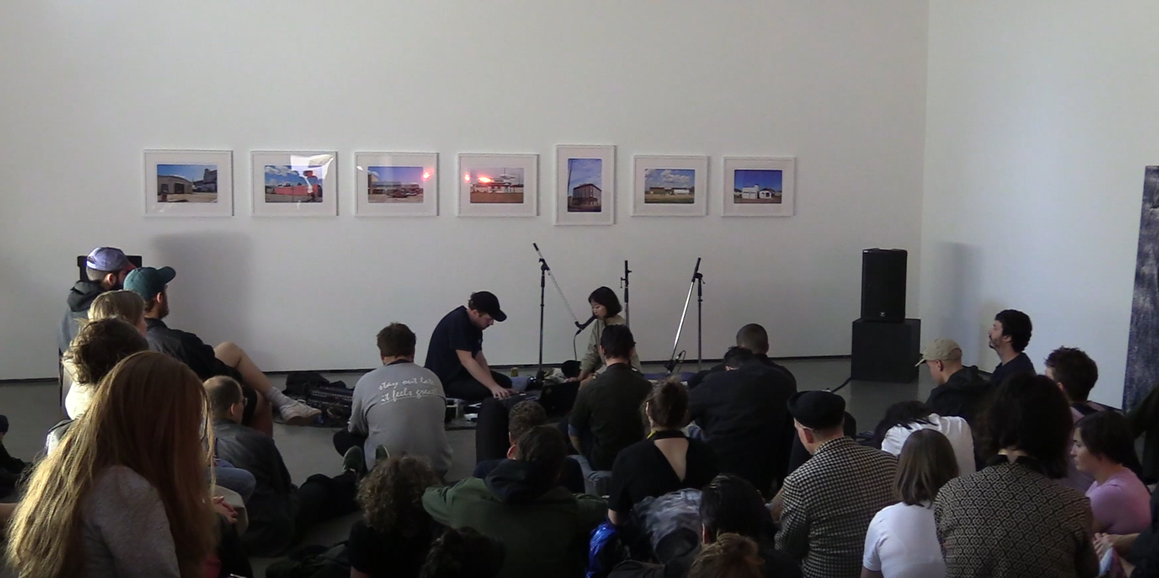 An image of a drumset and mikes being set up in front of a gallery wall. Seven landscape photographs are installed on the wall. The audience sit on the floor and waiting for the performance to start. 