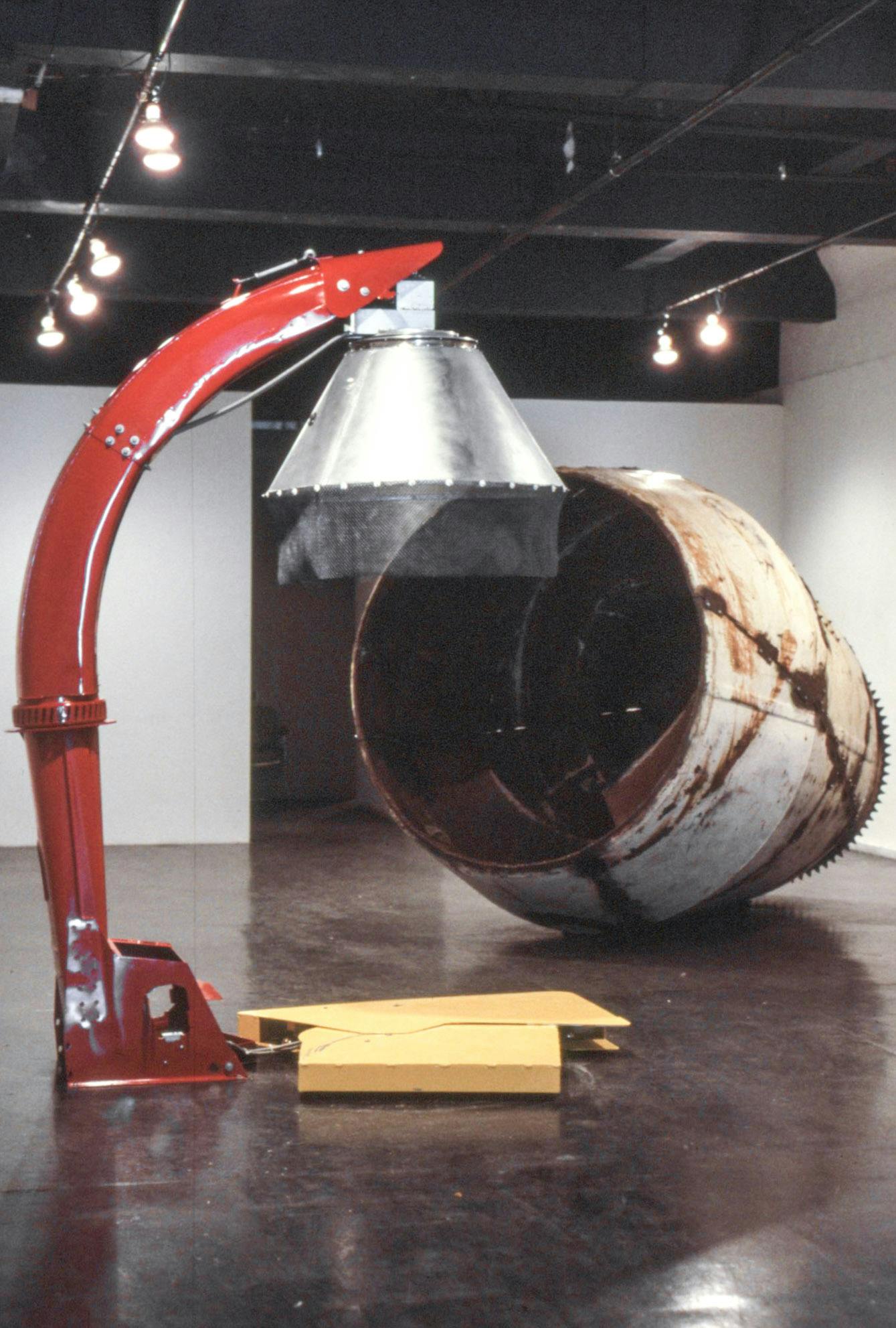 Two sculptures in a gallery space. The works are a large, rusty cement mixer and a bright red industrial silage spout.