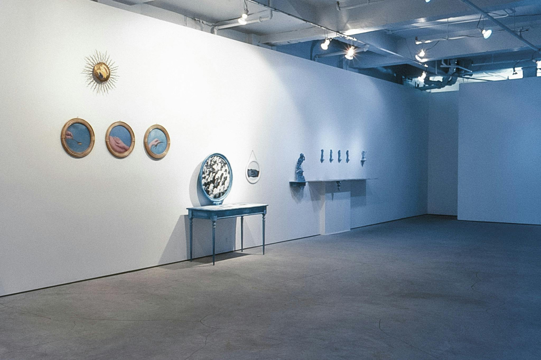 Artworks against and on the wall of a gallery. The works are mostly blue and include a trio of photos of hands on a blue background in gold frames, a blue vanity, and a row of small blue sculptures. 