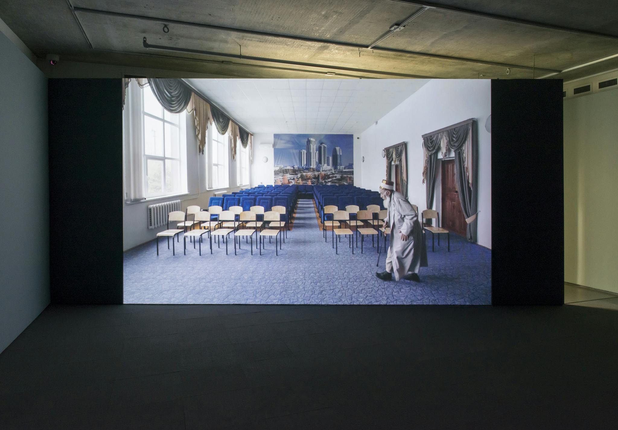 A large screen is installed in a darkened gallery displaying a projected single-channel video work. In the video, an older person with a white beard walks into a room filled with rows of chairs.