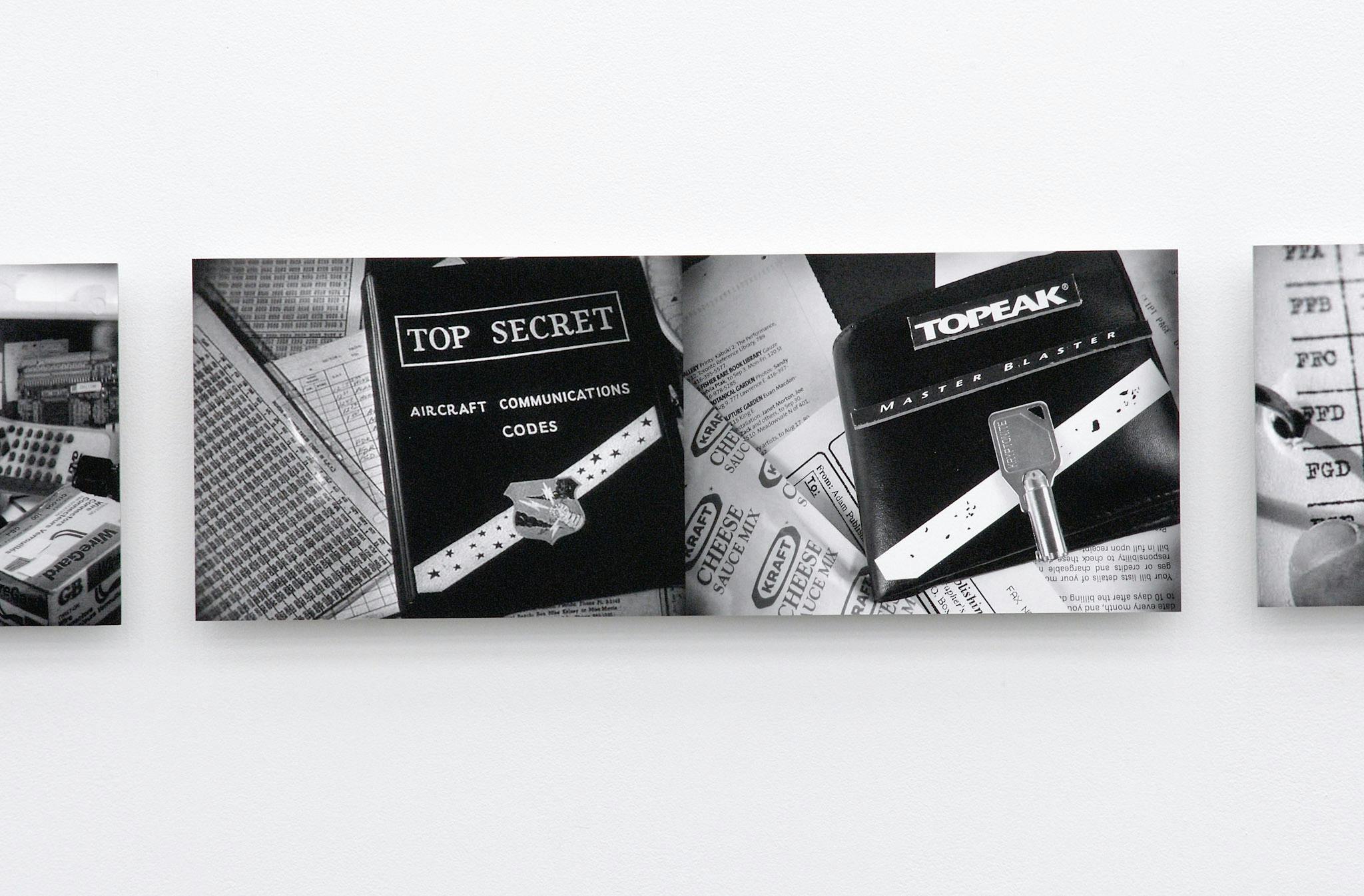 A pair of black and white photographs are installed on a gallery wall. The image on the left shows a book titled TOP SECRET. The next image mimics the left image with everyday objects.