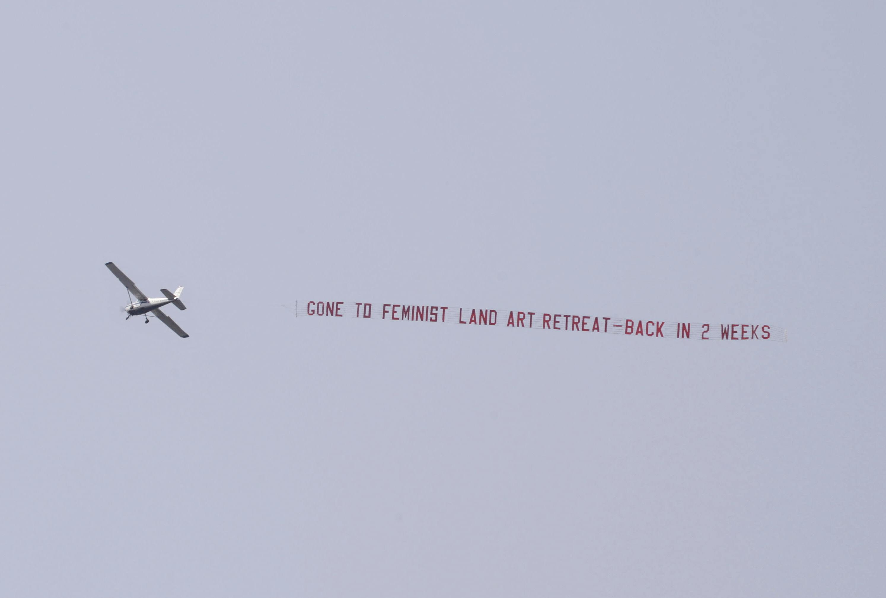 A small plane flying in the sky pulls a banner that reads in red text: “GONE TO FEMINIST LAND ART RETREAT- BACK IN 2 WEEKS”.