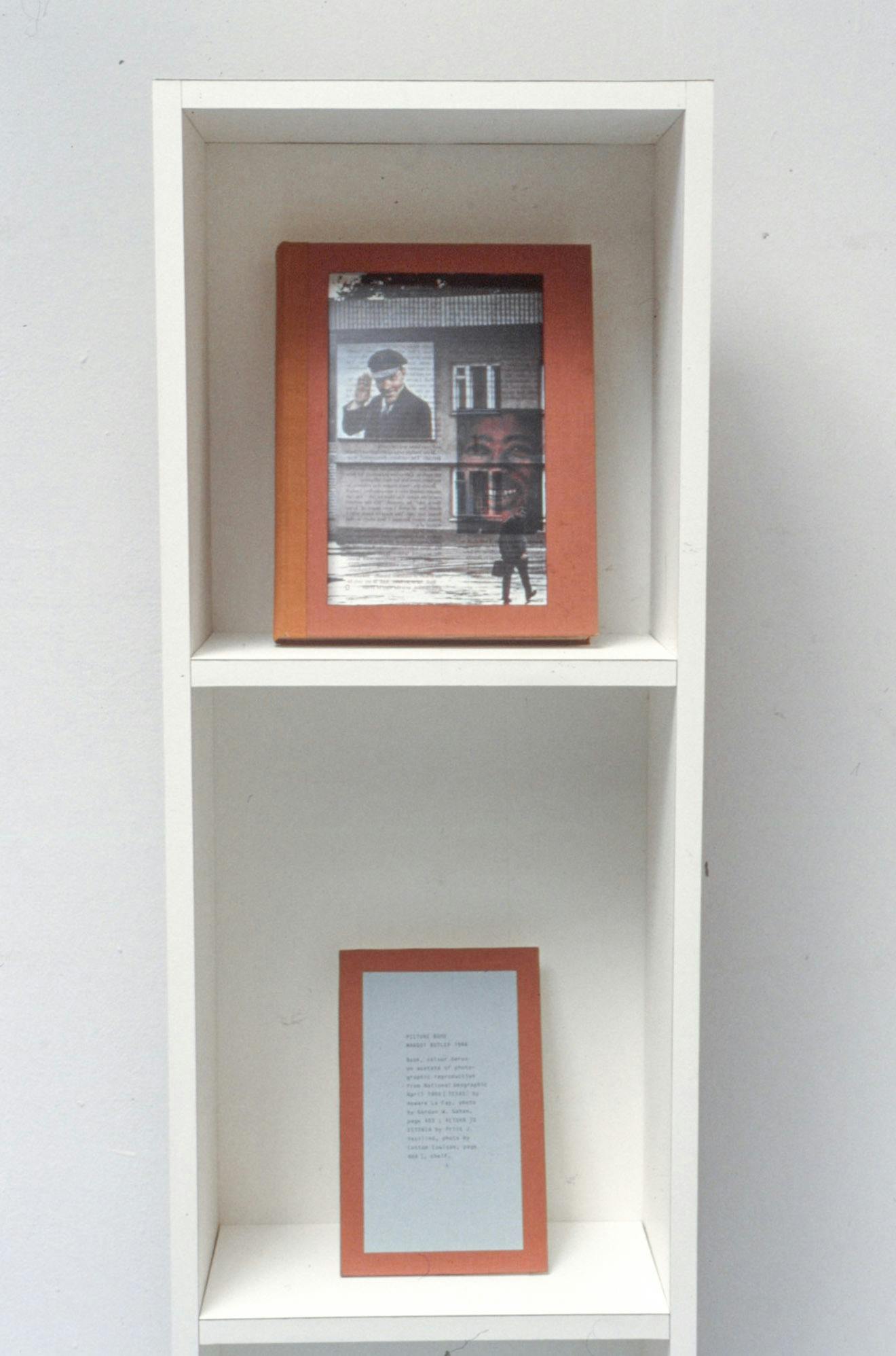 A closeup of a narrow white bookshelf against a wall. The top compartment shows a book with an orange cover, with a collage of faces and buildings. In the bottom there is a small poem in a wood frame.