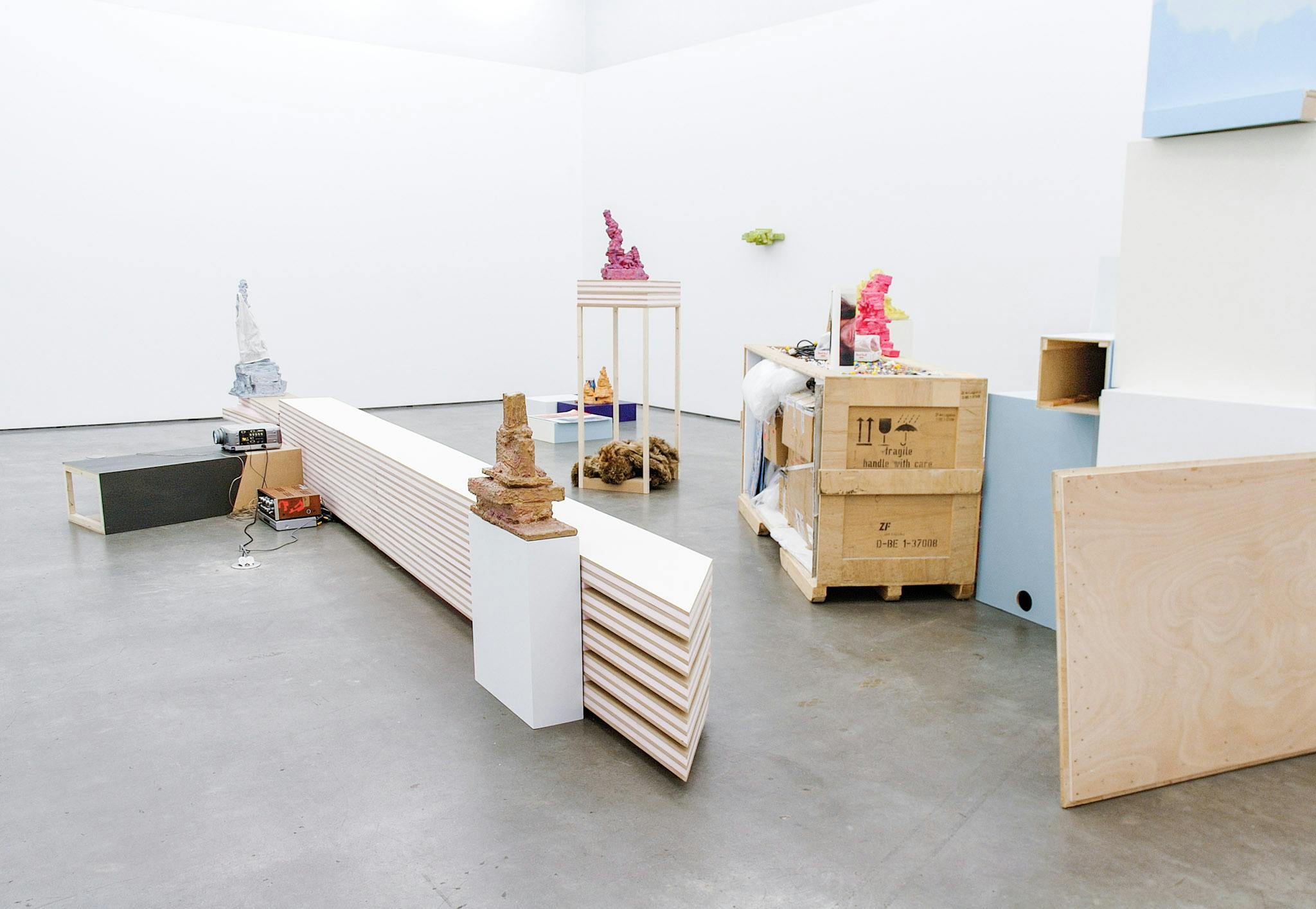 Various artworks in a gallery. The works are composed of wood, foam and paint. Some resemble shipping containers, some are small vertical constructions painted in bright colours like pink and green.