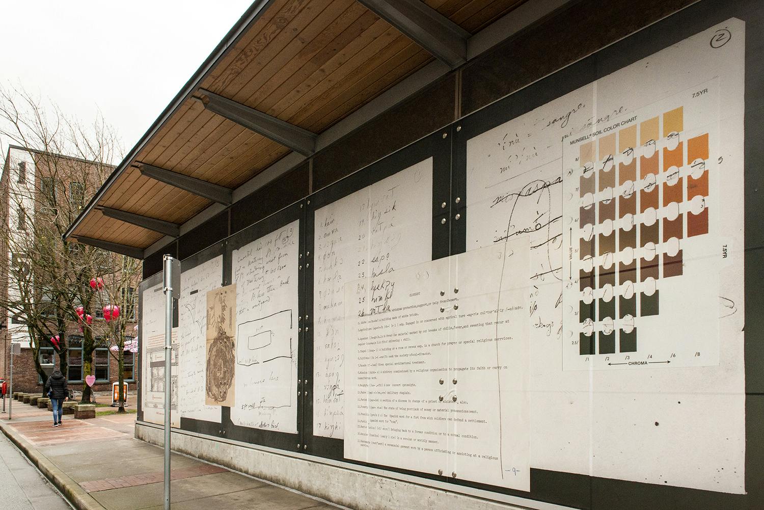 An exterior view of a train station displaying Sandoval’s work on the glass facade printed in vinyl. The work depicts a collage of scanned and enlarged archival documents, including written notes. 