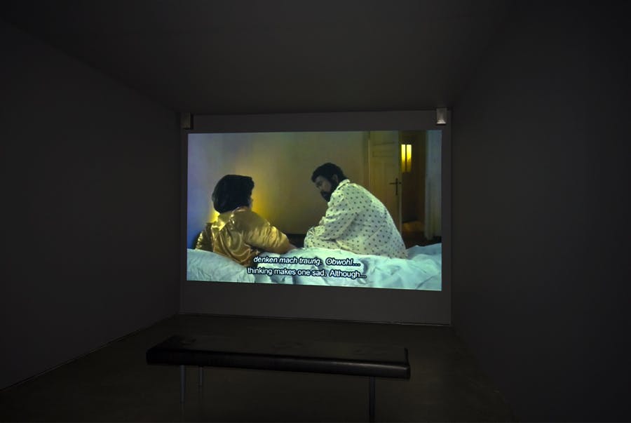 A projection on a screen in a dark room of a gallery. The screen shows two people sitting in a bed. There are subtitles in both English and German that reads “ thinking makes one sad. Although…”
