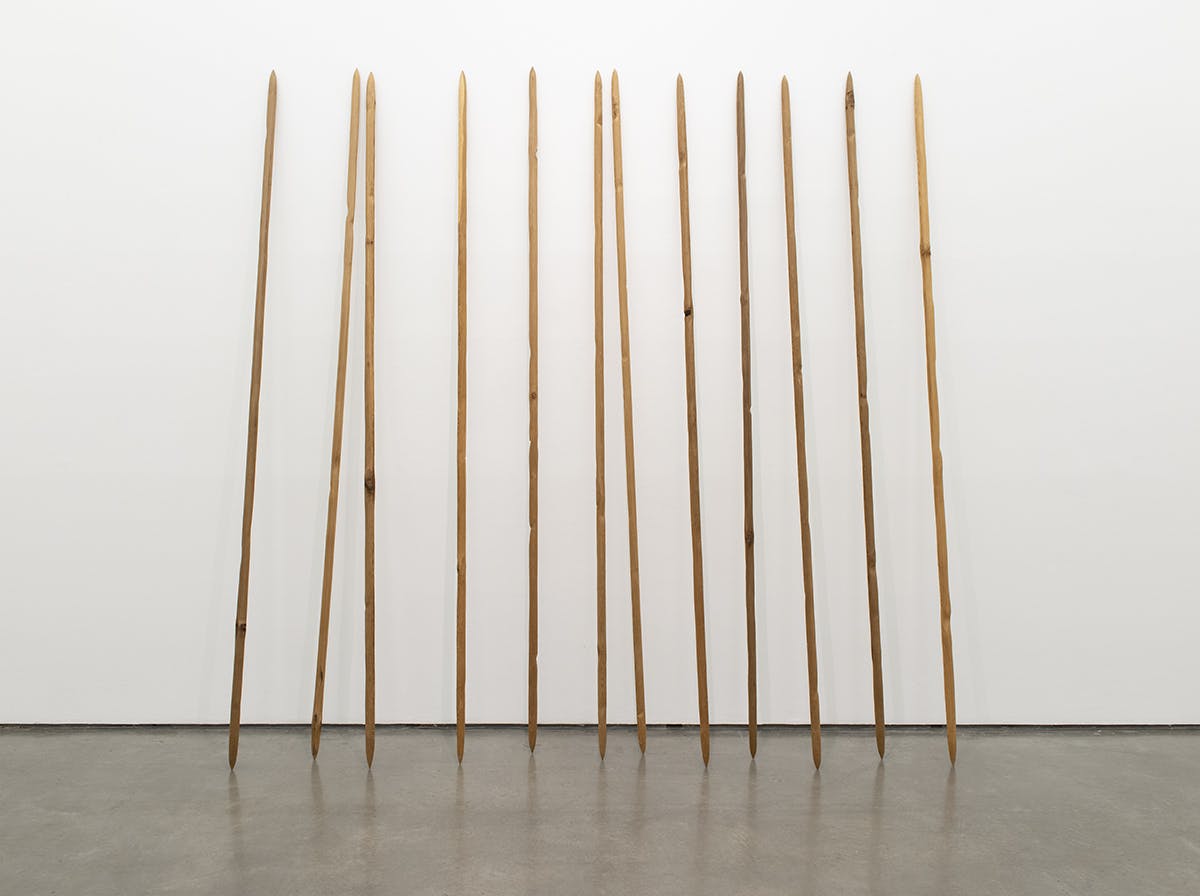 Twelve tall spear-shaped sculptures carved out of cedar lean against the gallery wall. They are all shaped in the same size and left unpainted. They stand vertically as if to make a fence. 