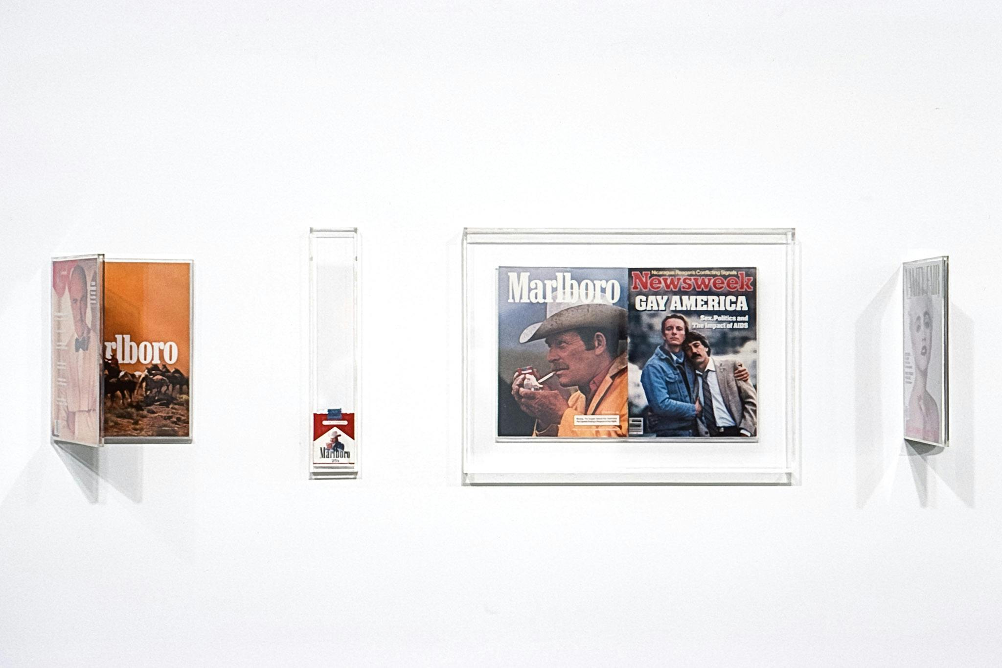 4 artworks in clear frames on a wall. They are a GQ magazine with a Marlboro ad, a pack of Marlboros, A Marlboro ad in a Newsweek magazine (with a cover reading: "Gay America"), and a Vanity Fair.